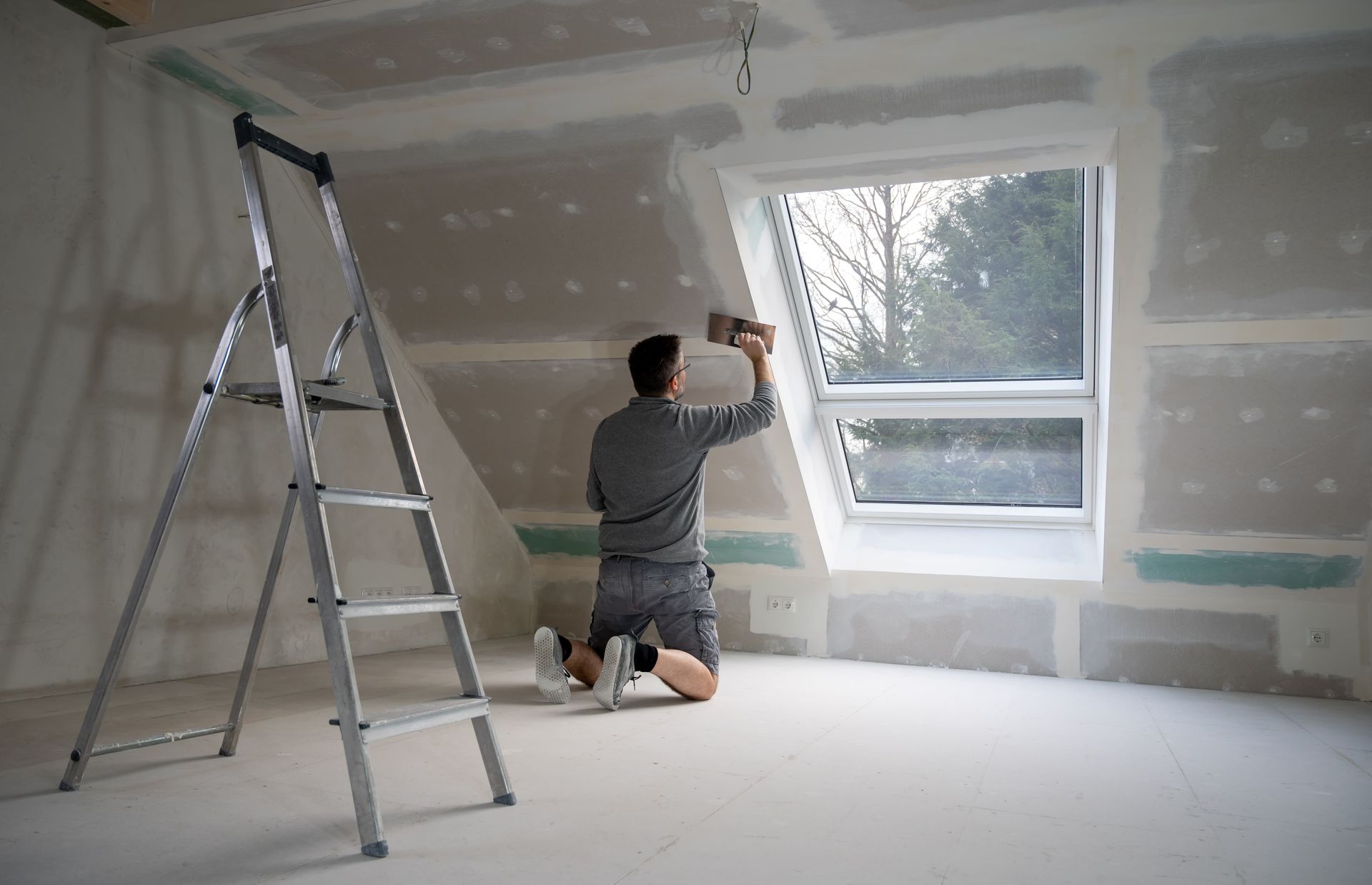 A man is kneeling down in a room with a ladder and a window.