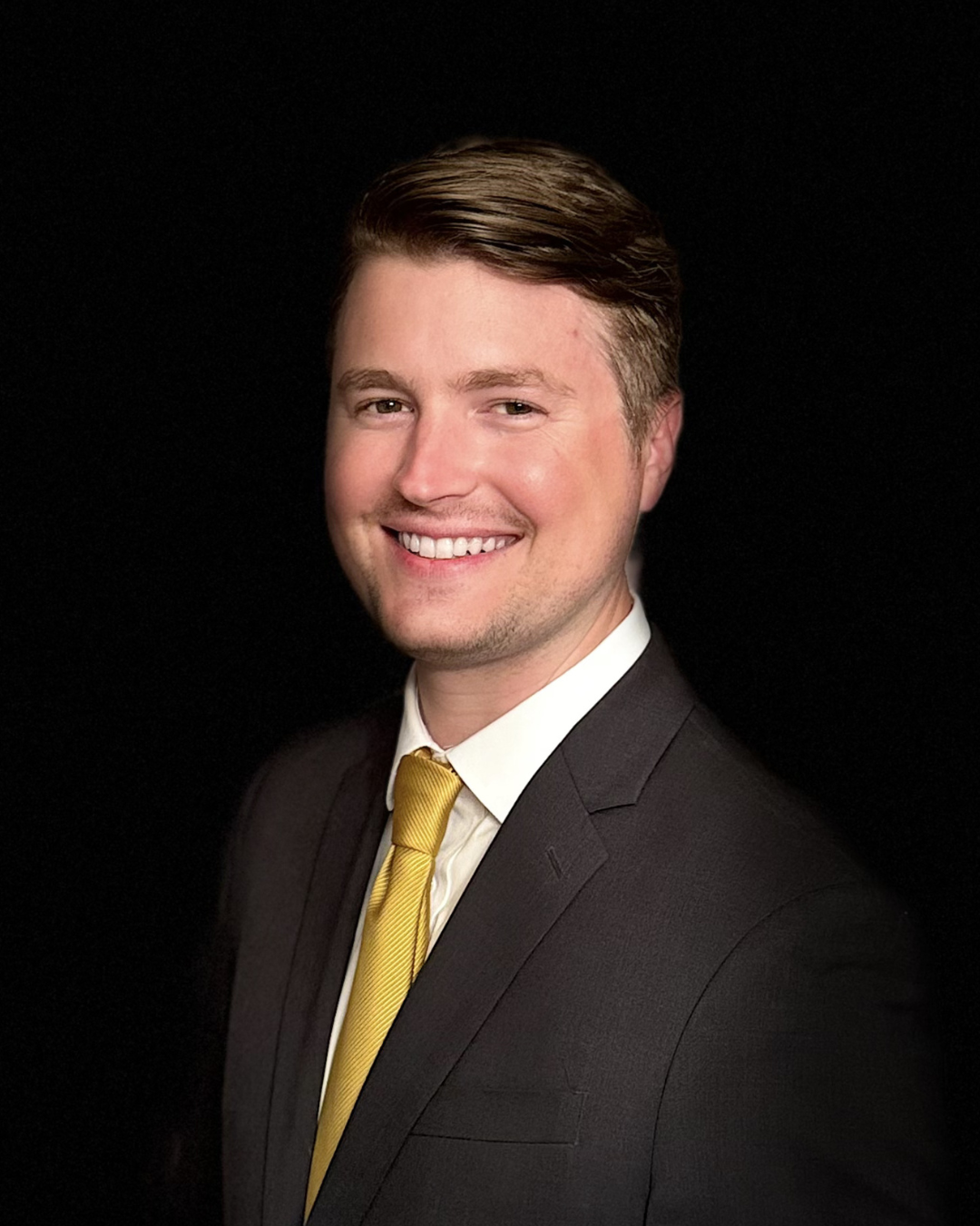 Man in navy suit, yellow tie, smiling. Blurry, bright background.