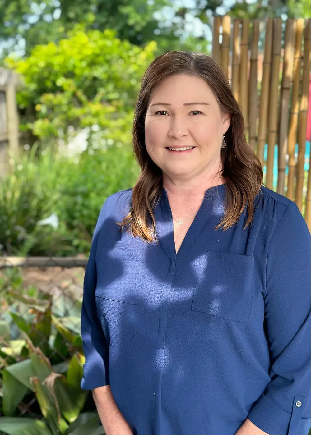 Woman with brown hair smiles, wearing a blue shirt, standing outdoors in front of greenery.