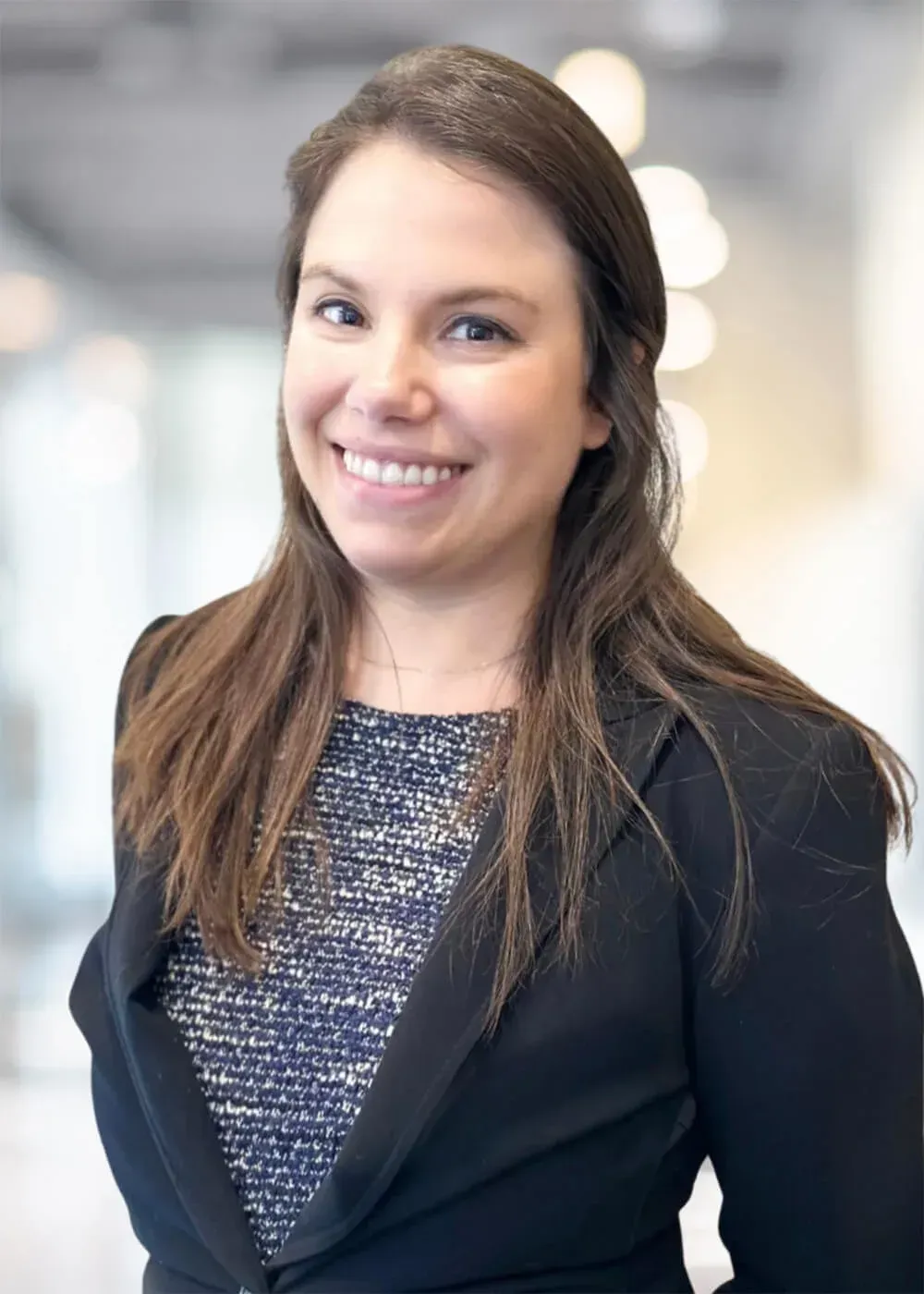 Woman in a blazer smiles, standing in a blurred office setting.
