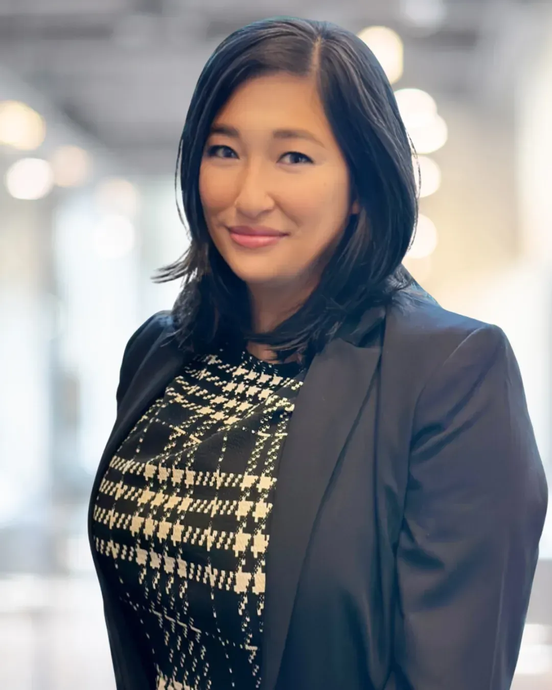 Woman in black blazer and patterned top, smiling in a blurred office setting.