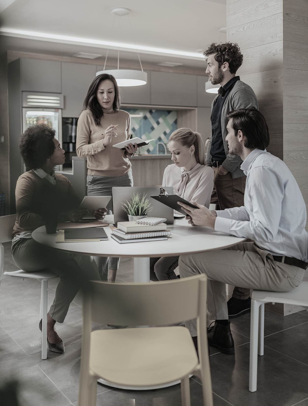 A business team meeting at a table, discussing work. A woman speaks, gesturing with a tablet. Others listen.