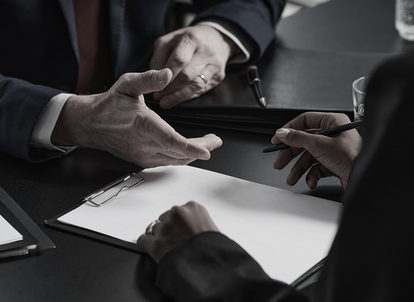 Two people at a table, one gesturing while the other writes on a clipboard.