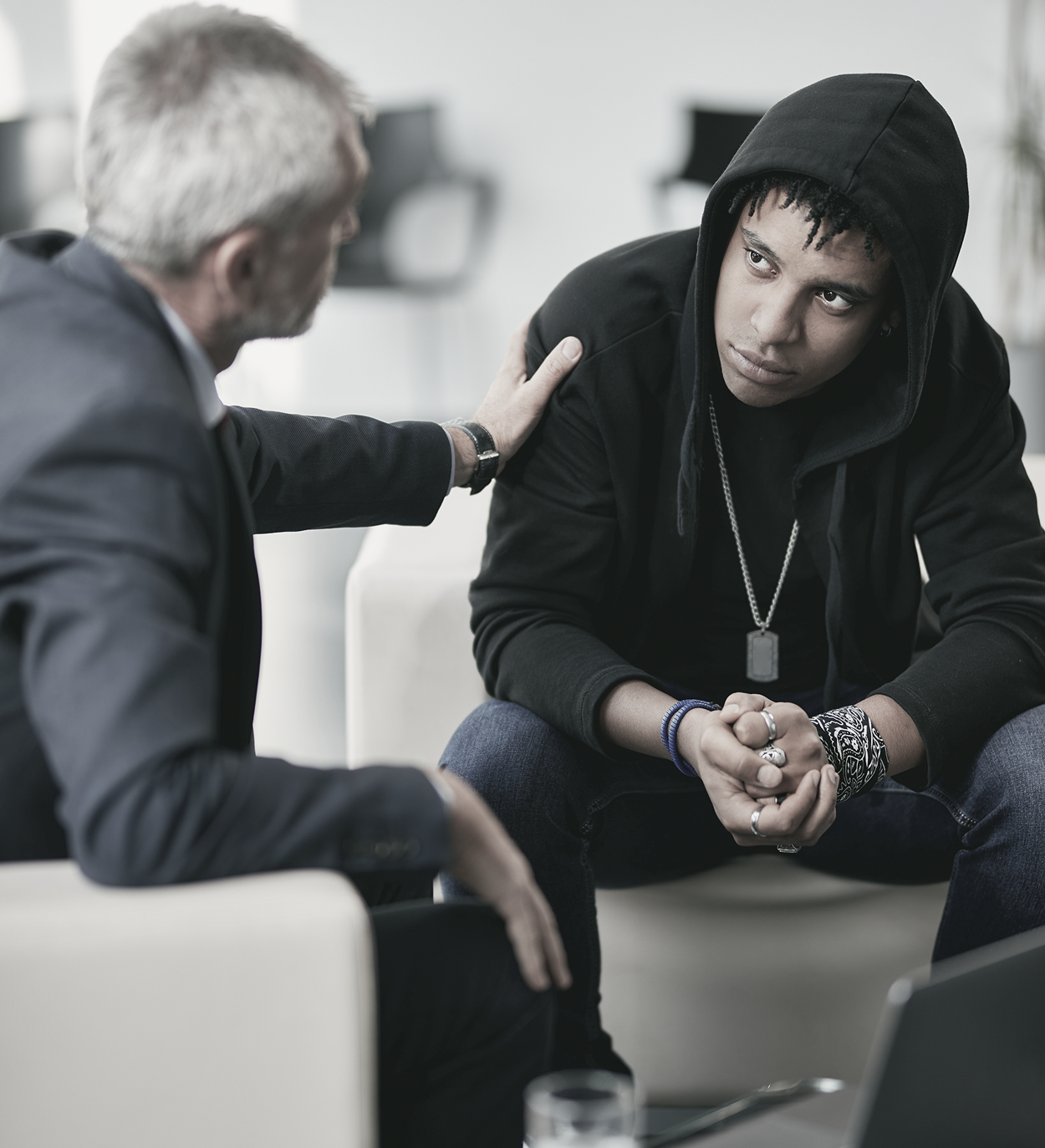 Man comforting a person wearing a hoodie, touching their shoulder. They sit in a well-lit space.