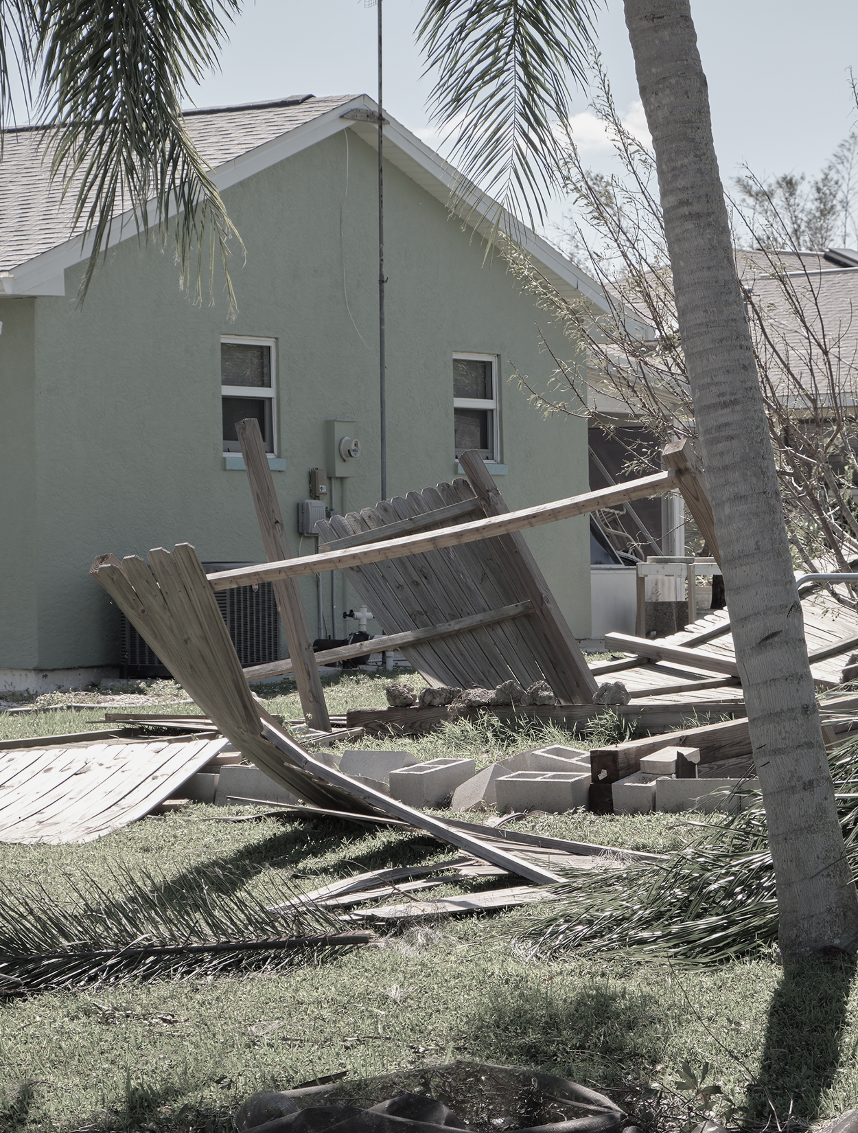 Damaged wooden fence leaning over, grass in front of a light green house. Palm tree on the right.