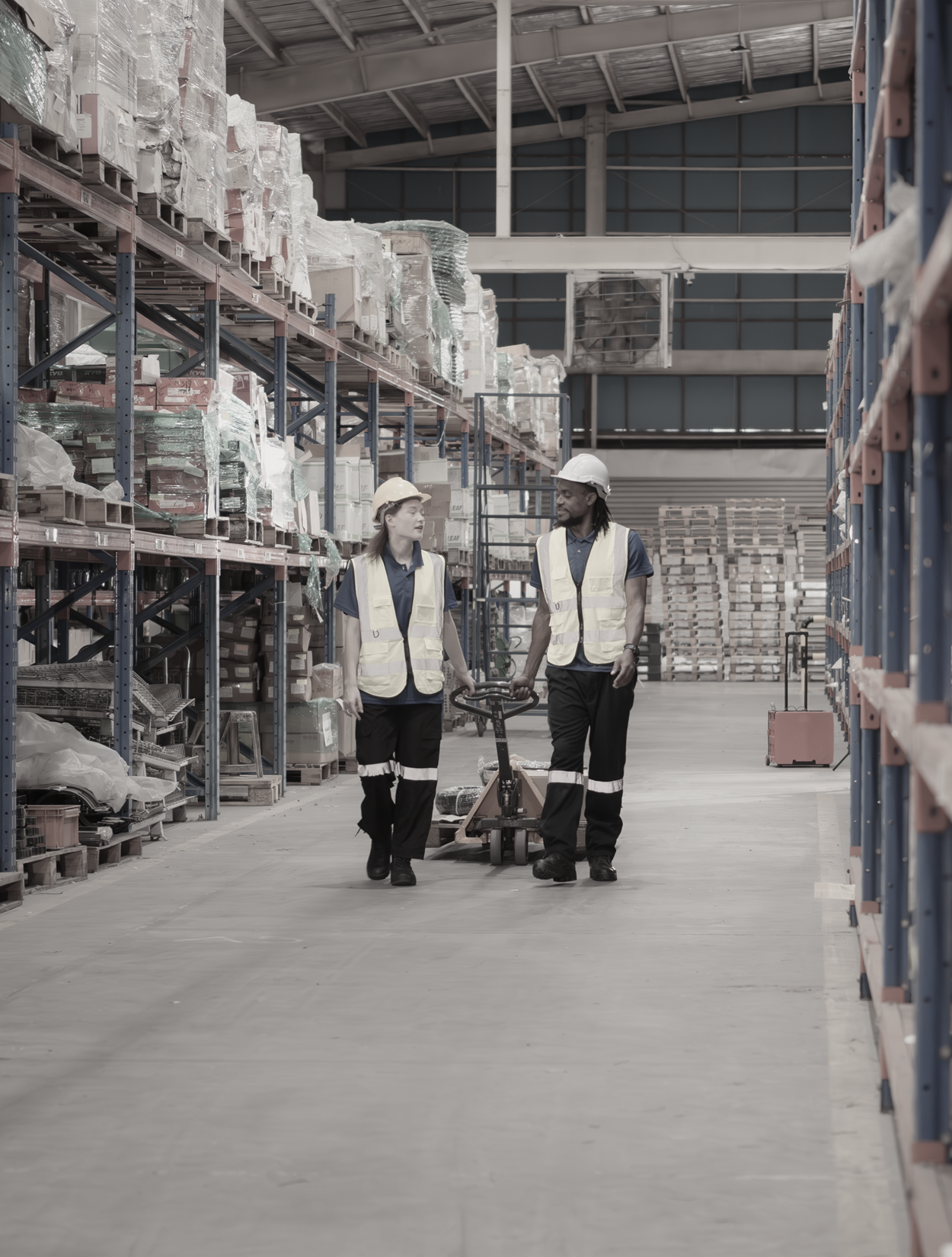 Two workers in a warehouse, walking and discussing near shelves stacked with goods.