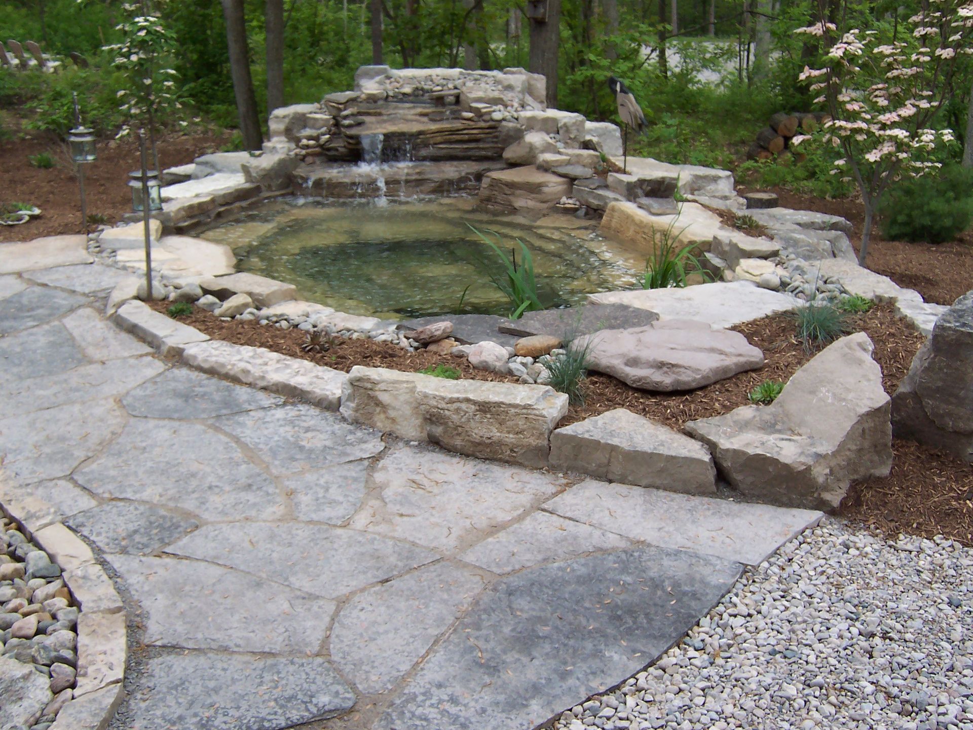 Stone pond with waterfall, surrounded by stone path and landscaping, in a garden.