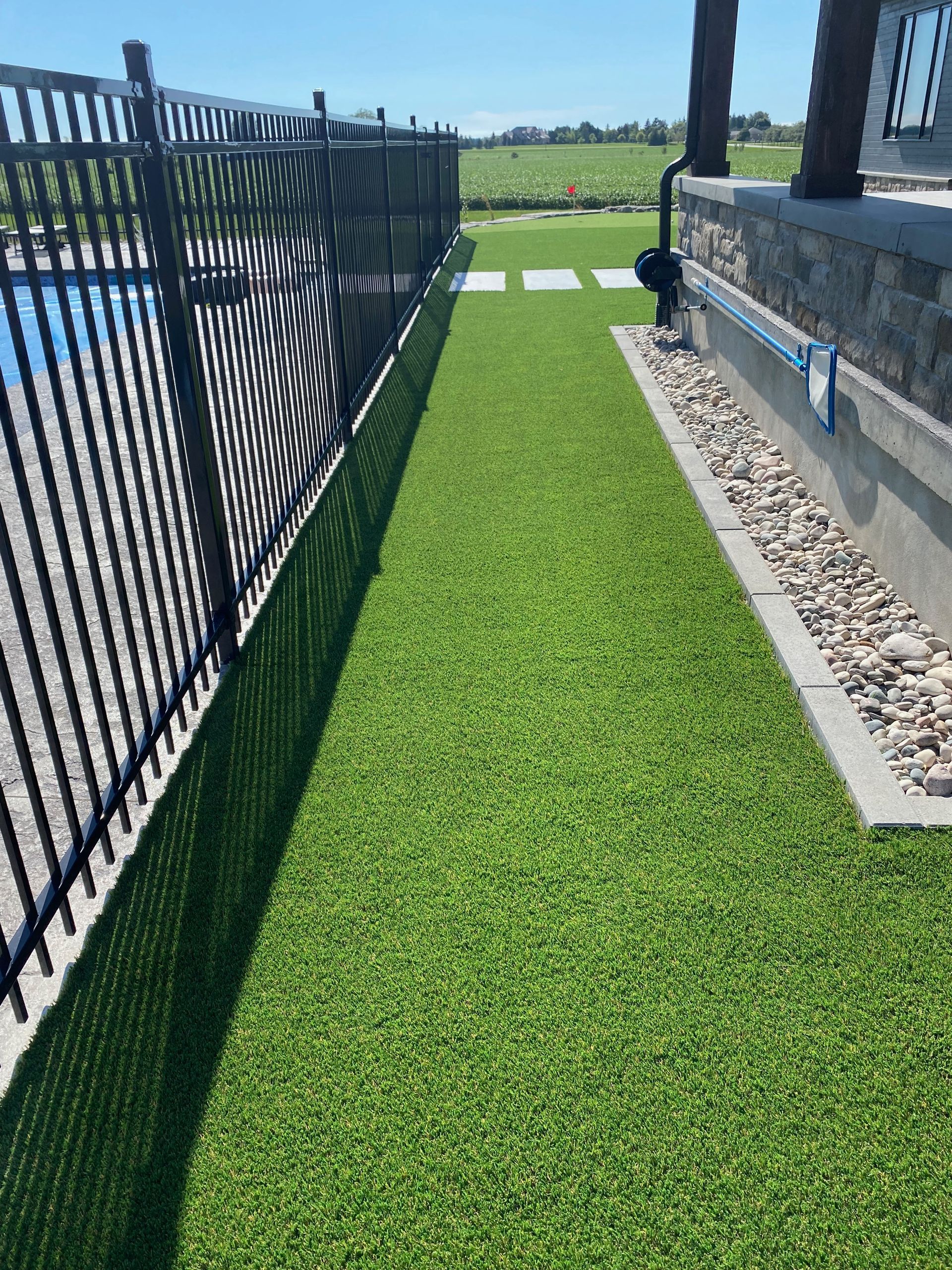 Narrow strip of artificial green grass next to a black fence and a building's foundation with rocks.