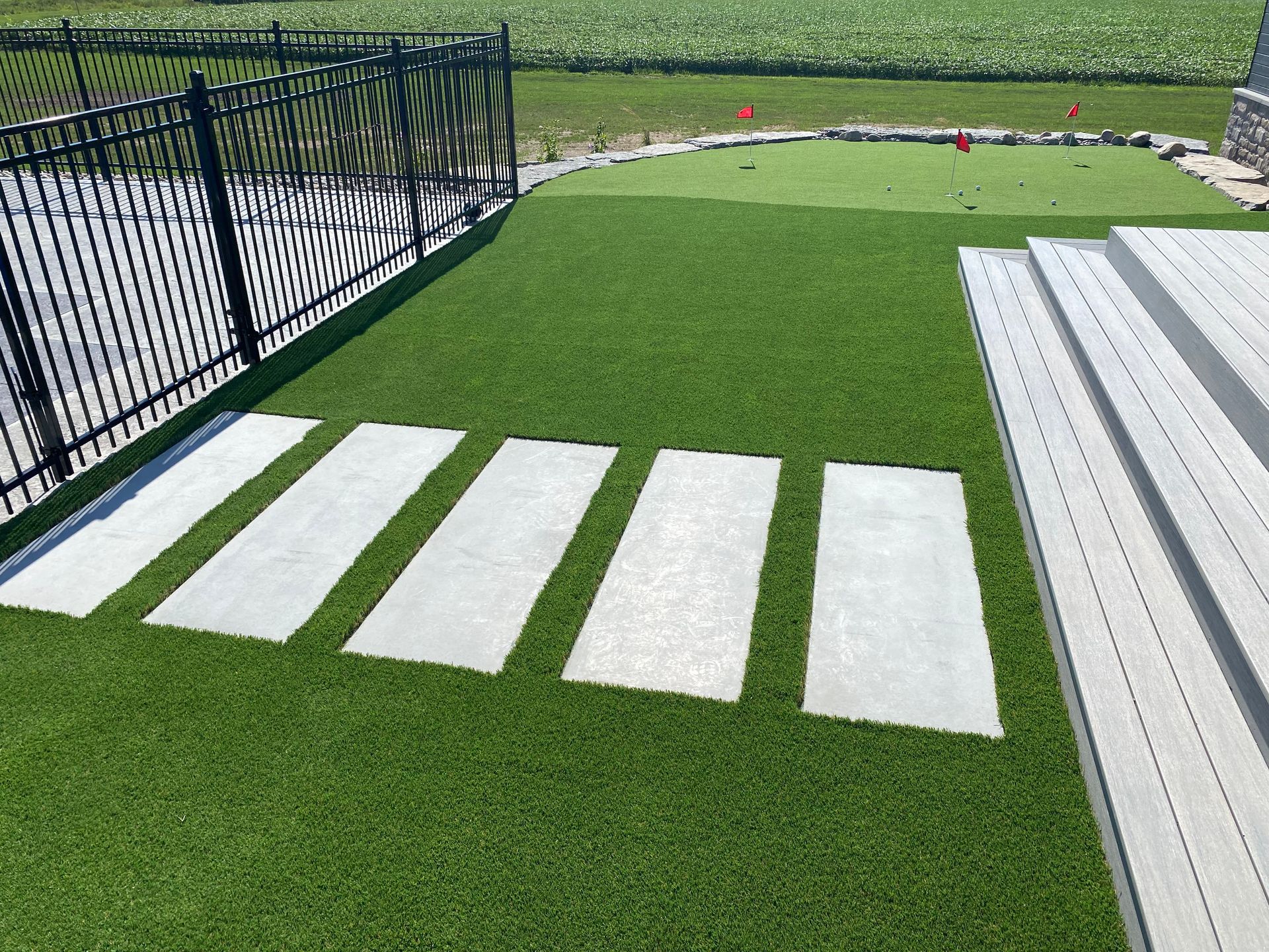 Rectangular stone stepping stones on green turf lead to a small putting green and deck. Black fence.