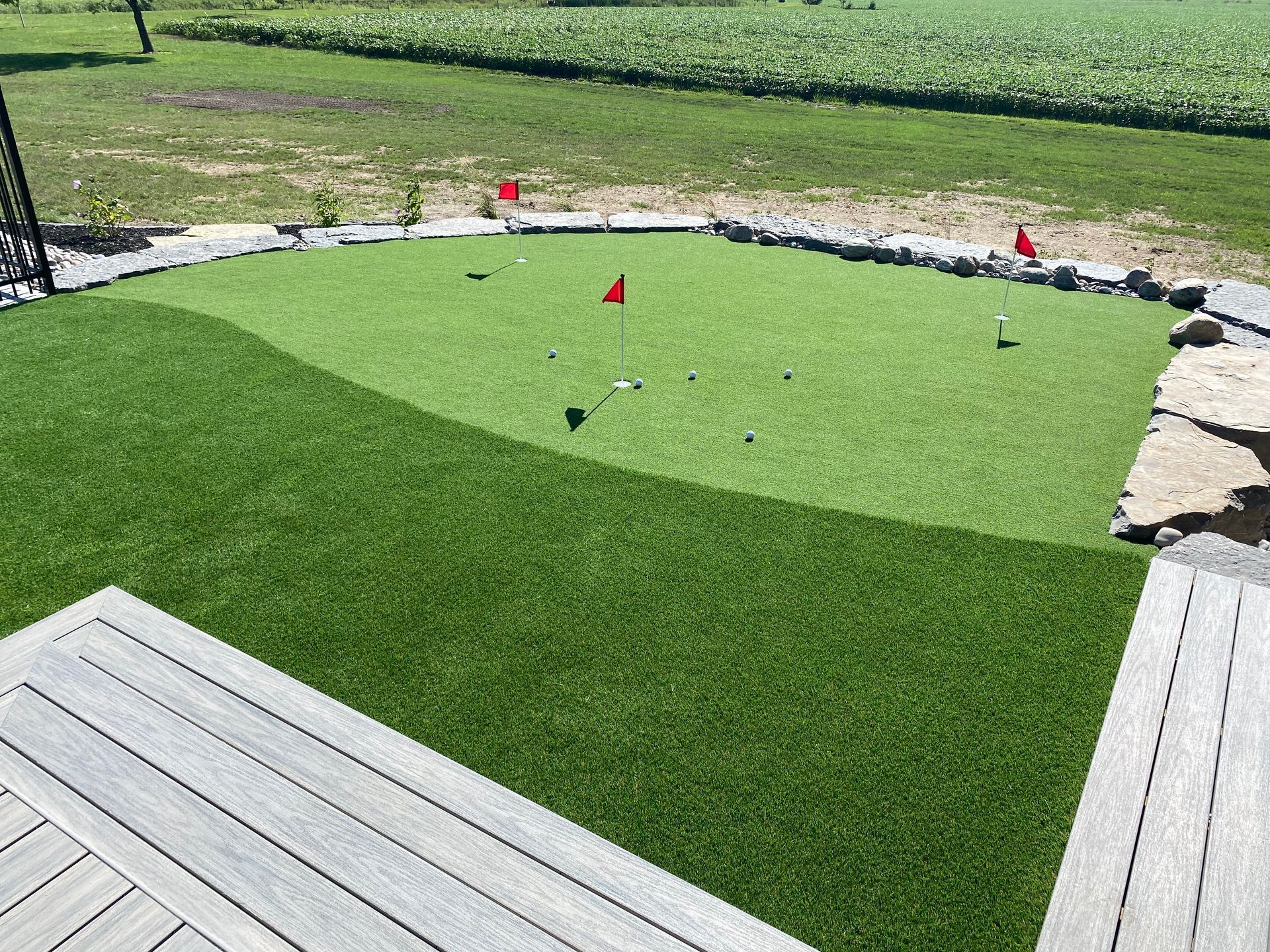 A backyard putting green with red flags, surrounded by rocks and a wood deck.
