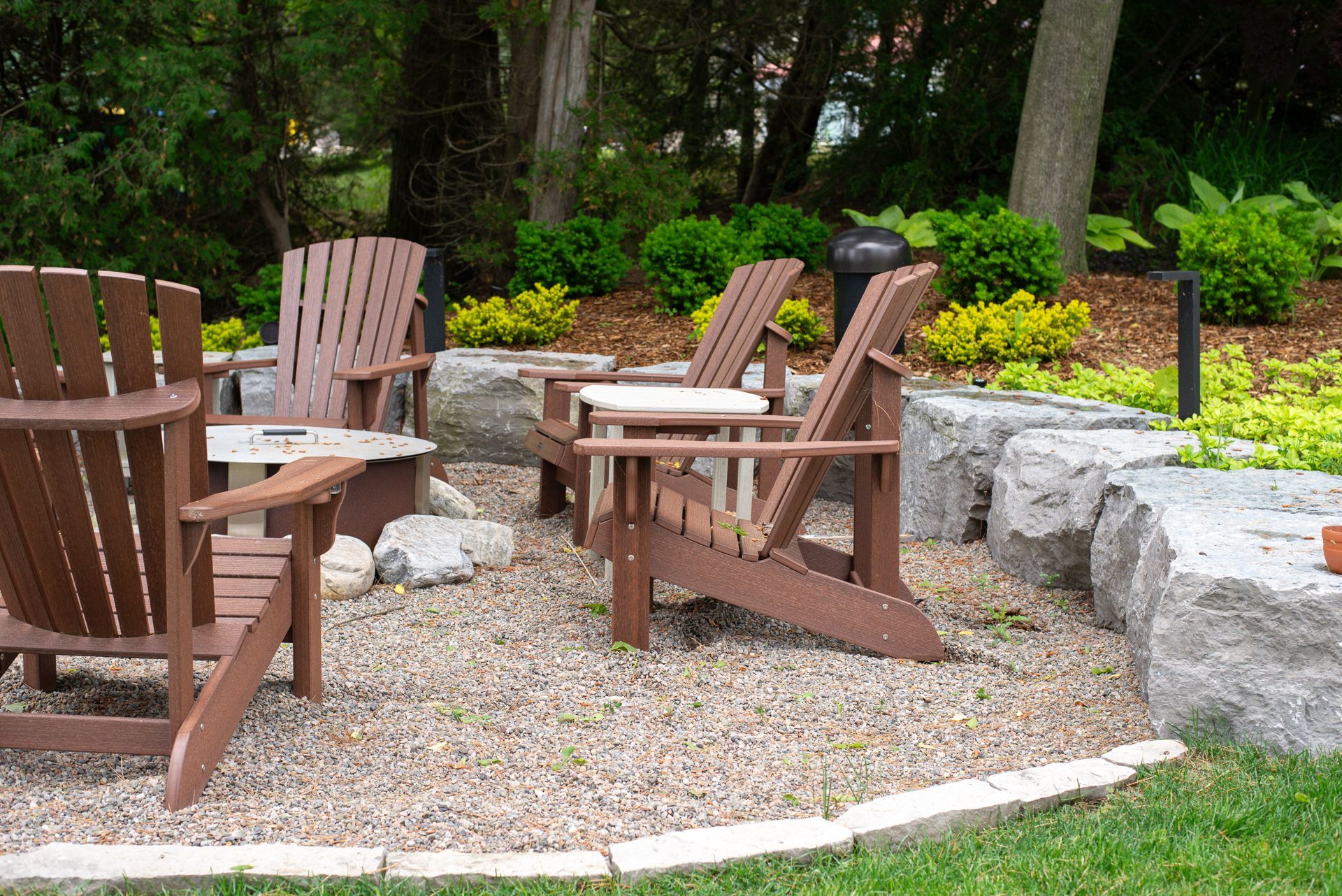 Brown Adirondack chairs around a fire pit in a gravel patio, with a rock wall and greenery in the background.
