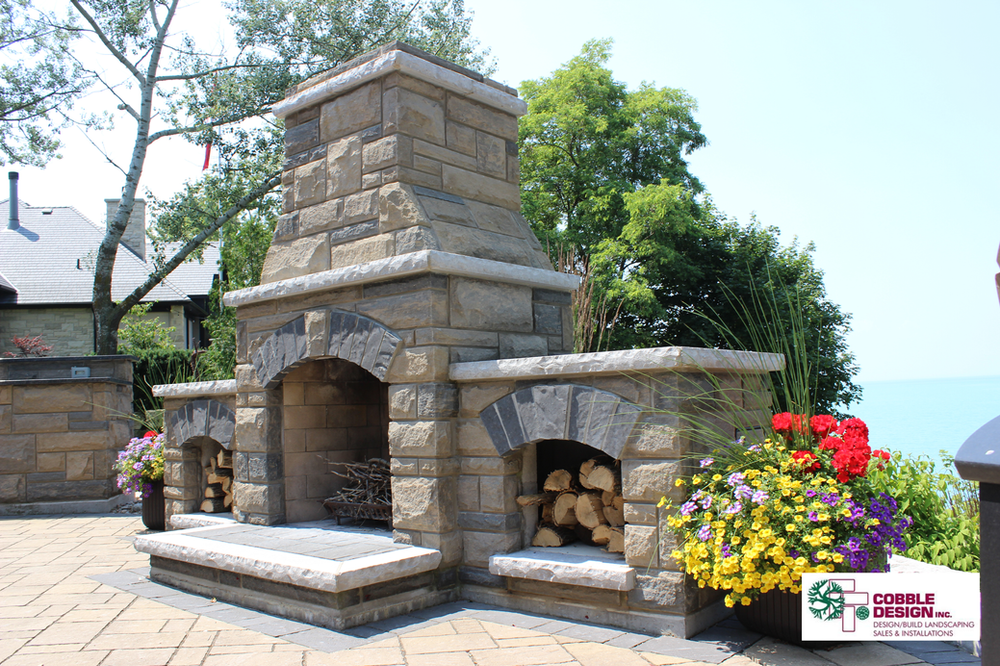 Stone outdoor fireplace with an arched opening, flanked by wood storage and flower pots, overlooking water.