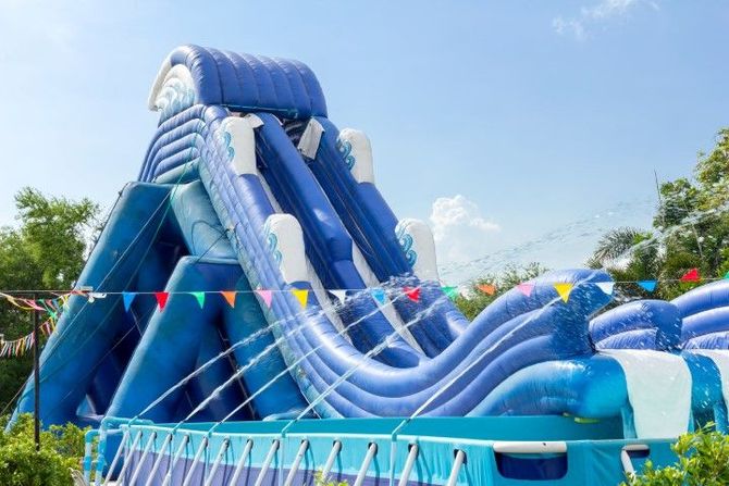 Large blue inflatable water slide at a sunny outdoor park with colorful pennant flags