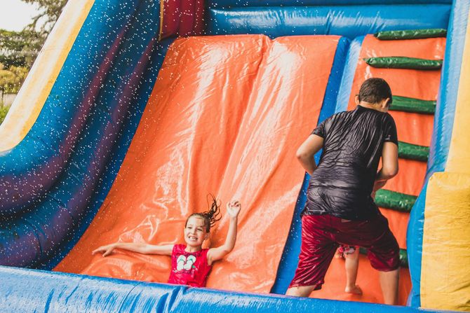 Child on inflatable slide with an adult nearby, in a colorful bounce house play area