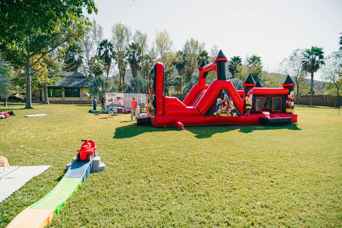 Large red inflatable obstacle course on a grassy park lawn, with a small slide in the foreground.