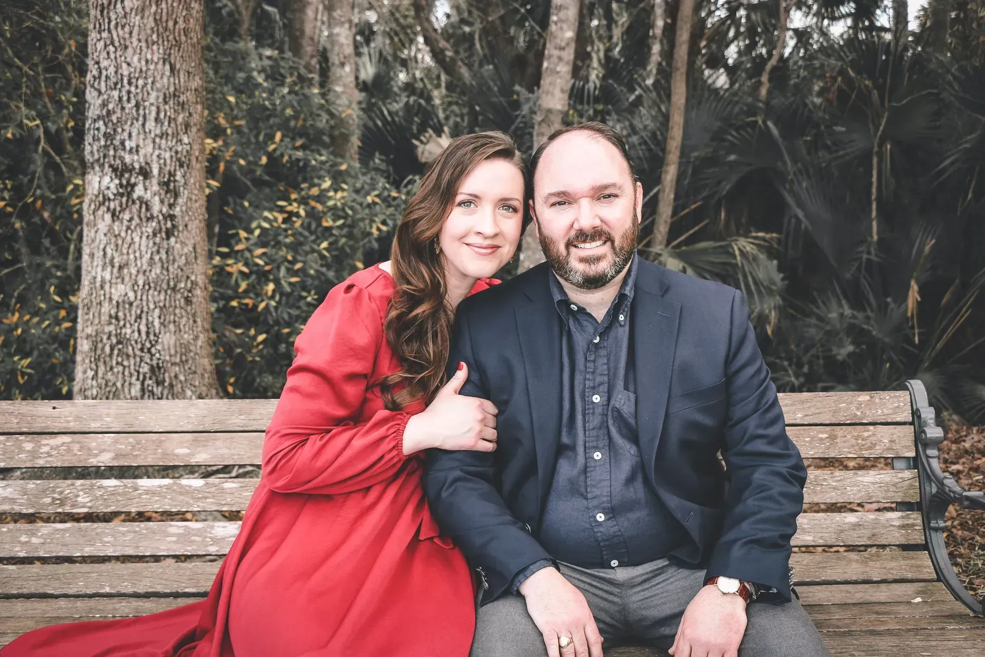 Couple sitting on a park bench, woman in a red coat leaning on a man in a dark suit, trees behind them