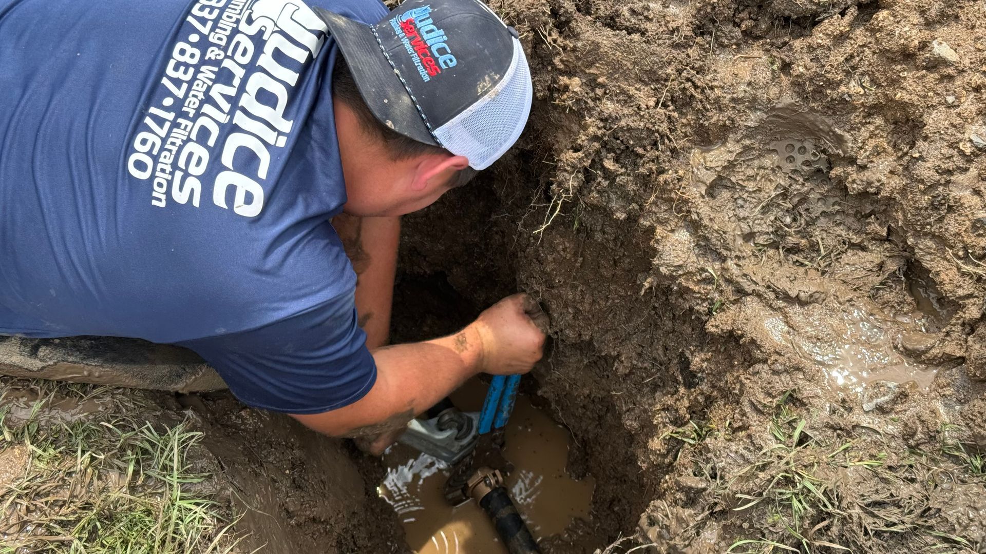 Plumber in a muddy ditch, fixing a water pipe with a wrench. The man wears a blue shirt and hat.