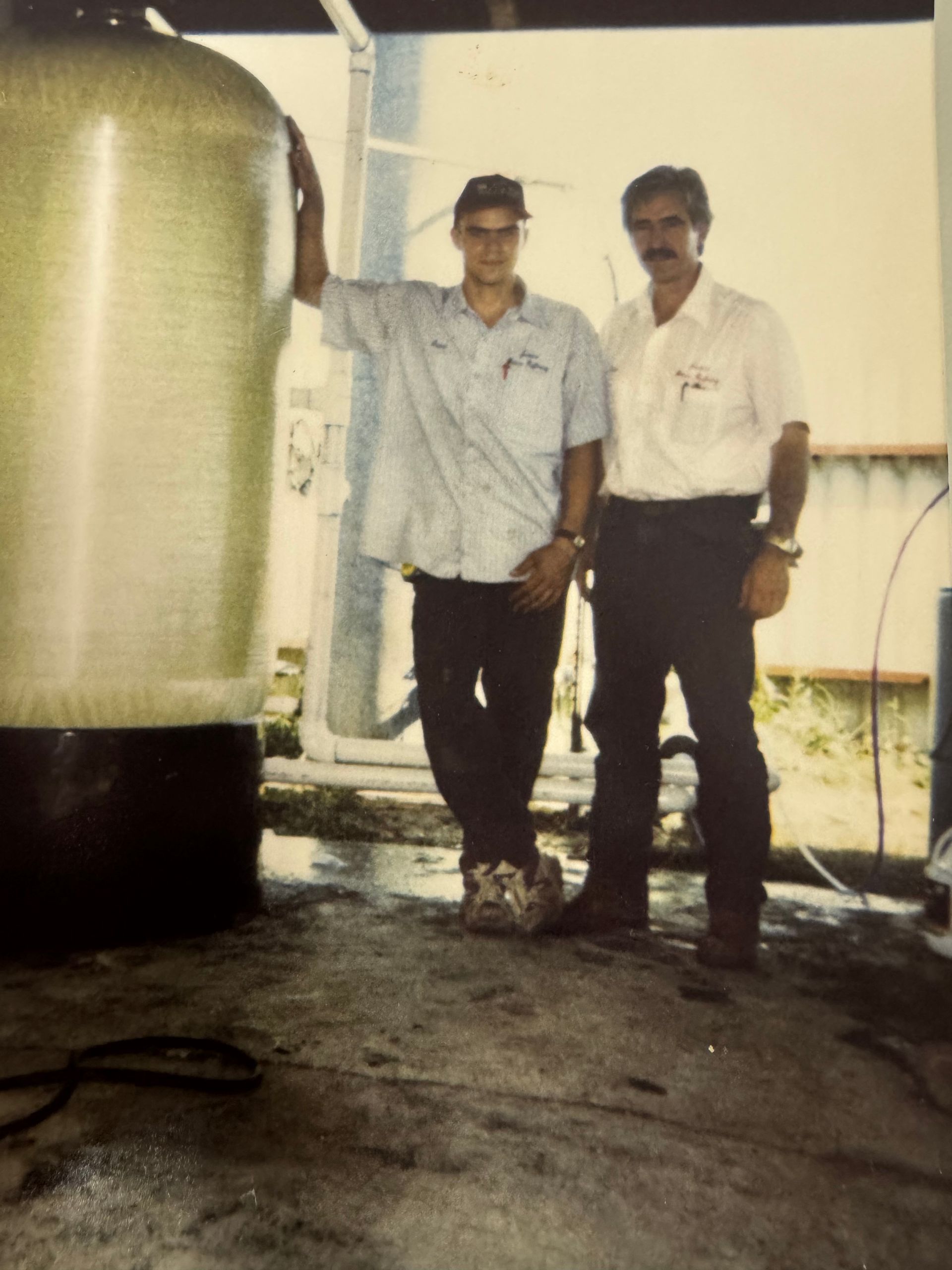 Two men standing next to a large water tank, one man leans on it. Outdoor setting, concrete floor.