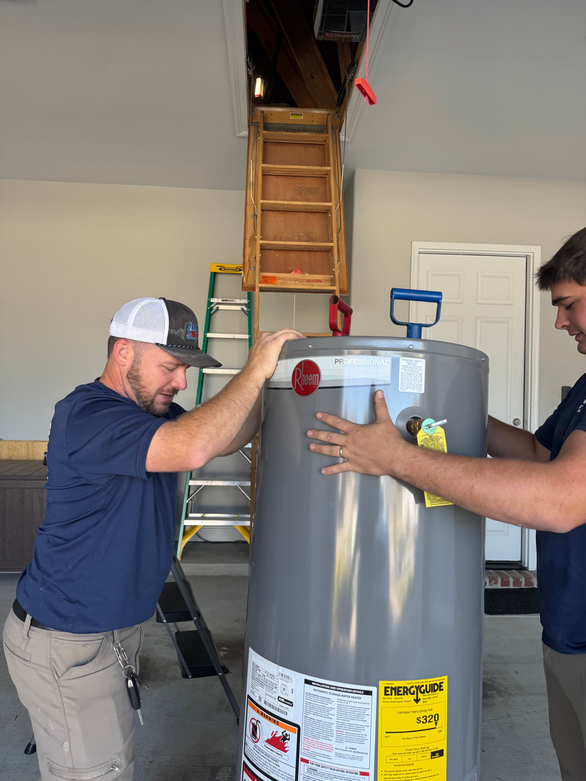 Two people moving a gray water heater in a garage. One man is in a hat and the other is beside him.