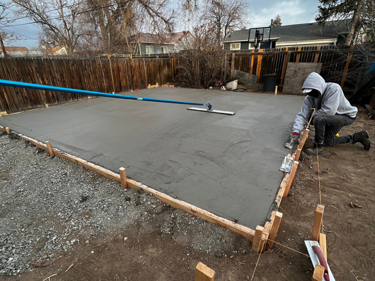 A person in a hooded sweatshirt kneels to finish smoothing a wet concrete slab framed by wooden boards in a backyard.