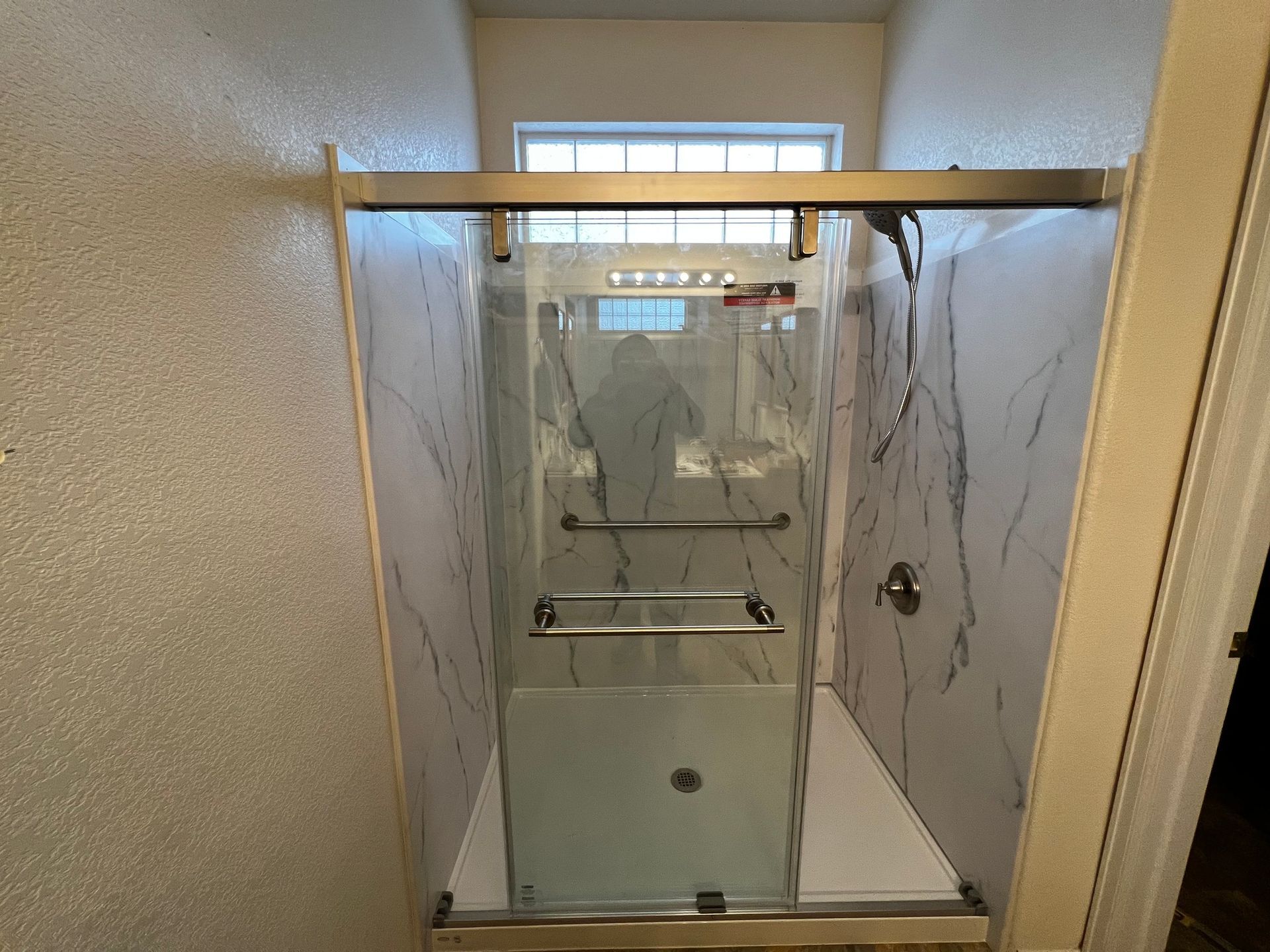 A walk-in shower with white marble-patterned walls, a glass sliding door, and silver grab bars, set in a tiled alcove.