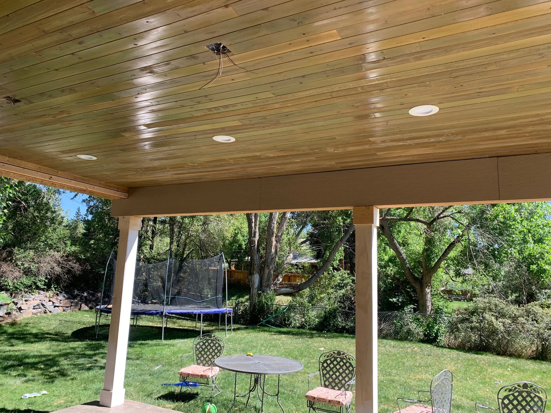 A covered patio with a wooden tongue-and-groove ceiling, recessed lighting, and support columns overlooking a green yard.