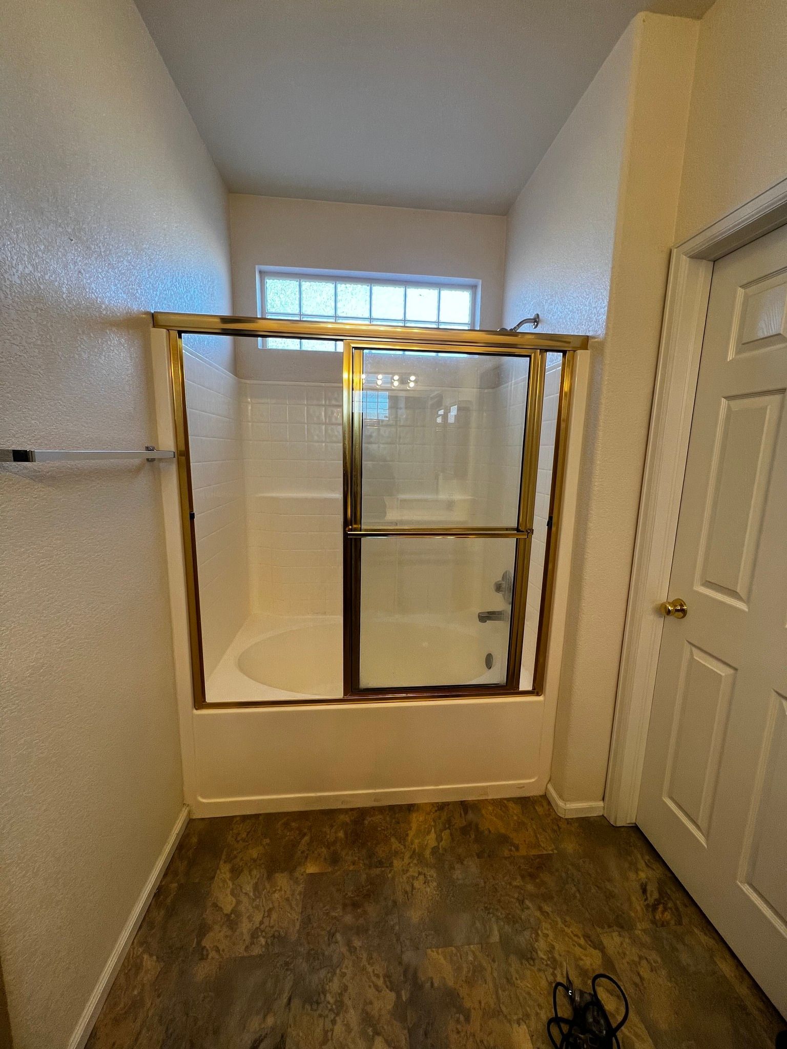 A bathroom bathtub with a sliding glass door and gold-colored frame, set against white walls and a dark tile floor.