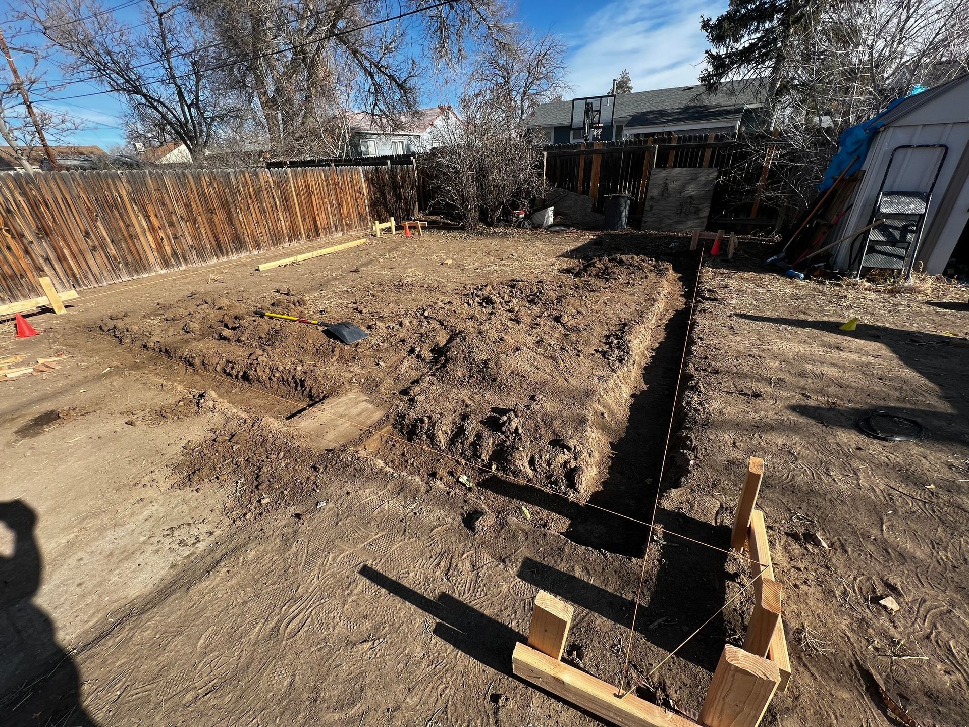 A backyard excavation showing trenches and wooden formwork prepared for a new foundation on a sunny day.