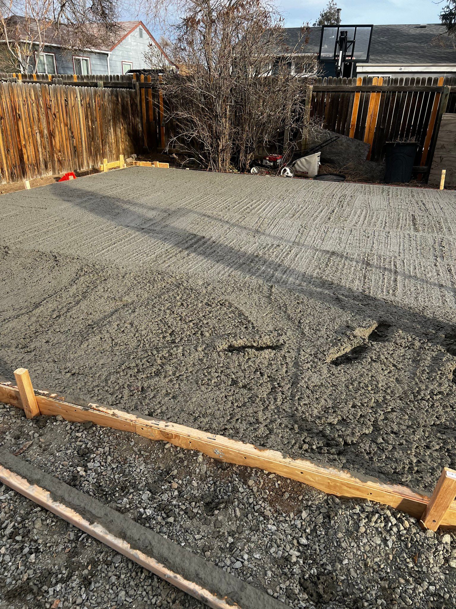 A level base of crushed gravel prepared for a concrete pour, outlined by wooden formwork boards in a residential backyard.