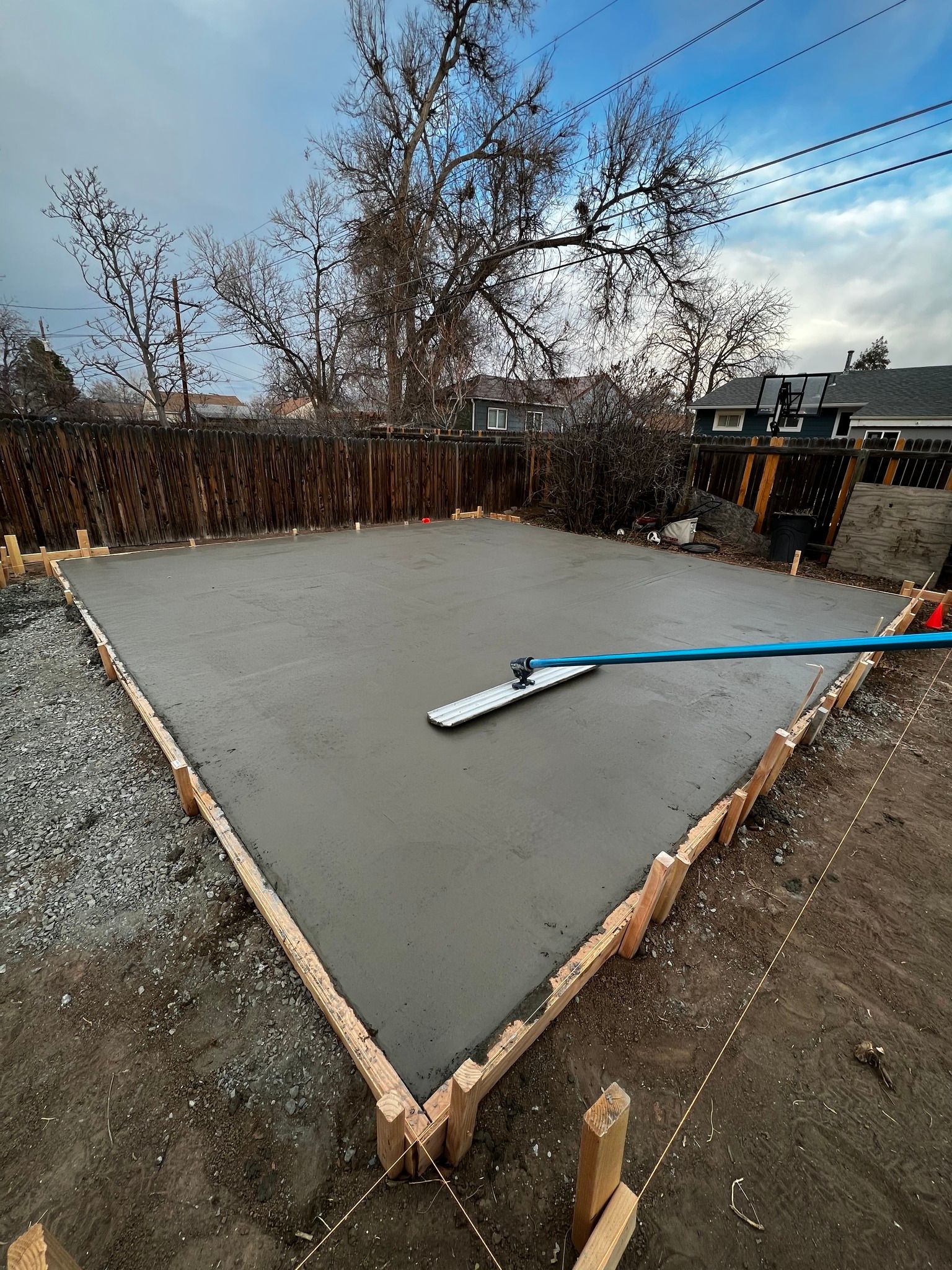 Freshly poured concrete slab in a residential backyard, framed by wood forms with a bull float tool resting on the surface.