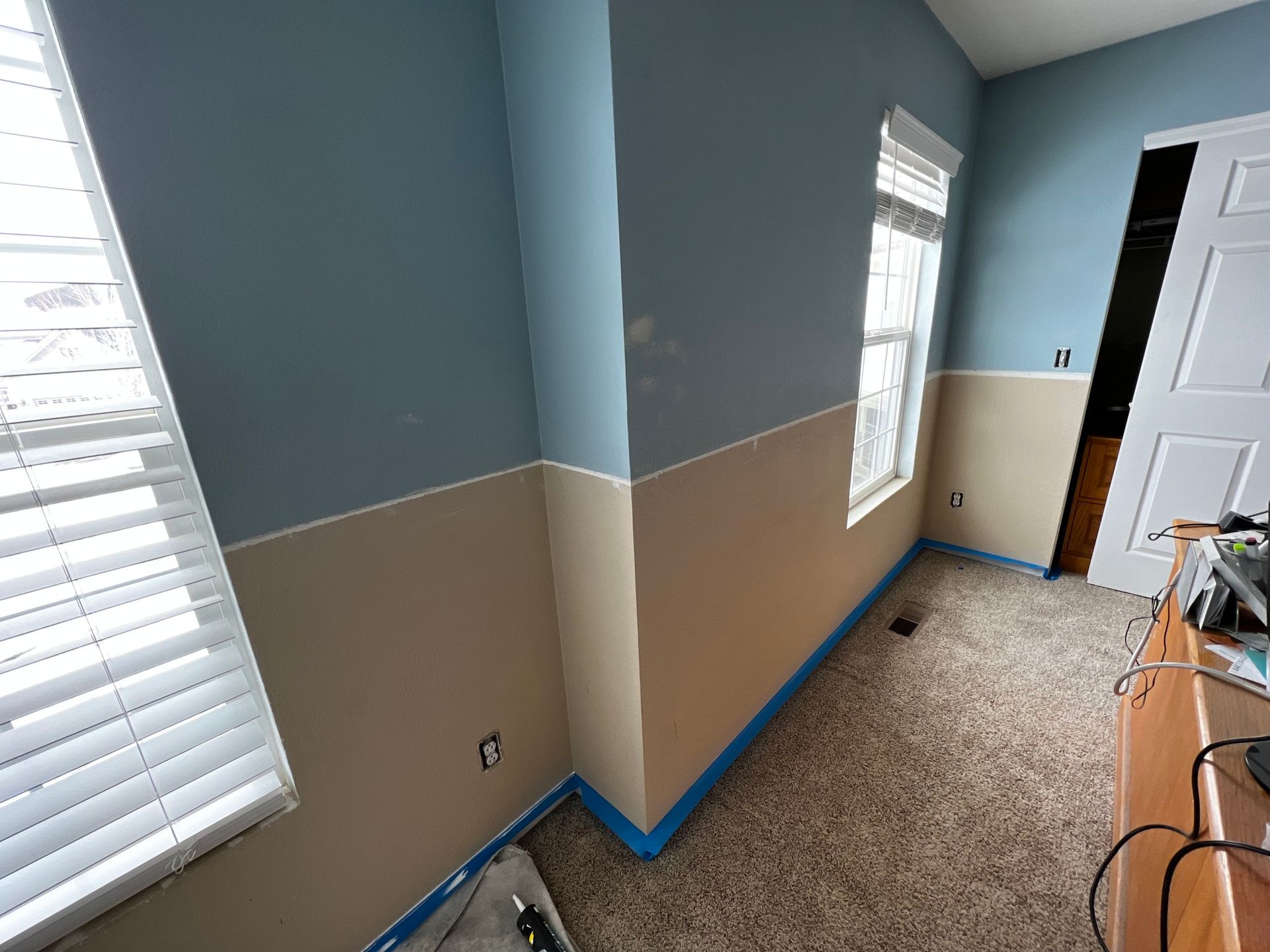 A room under renovation with blue and beige two-tone walls, blue painter's tape on baseboards, and windows with blinds.