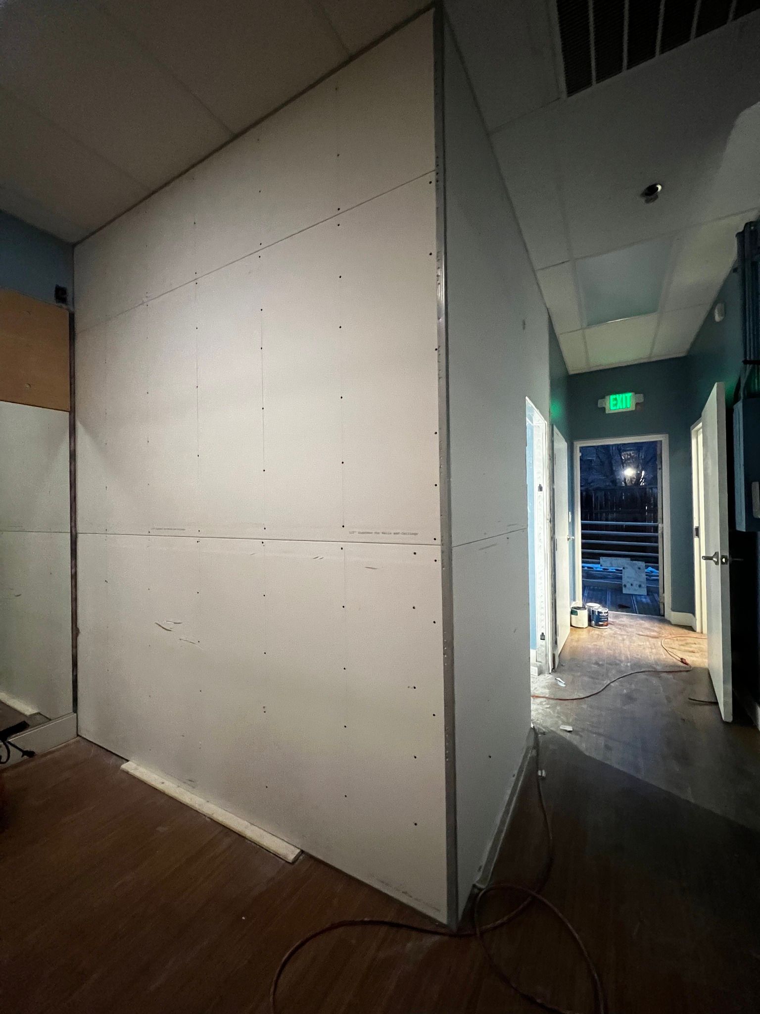 An unfinished wall covered in drywall panels stands in a room, leading to a hallway with an open exit door.