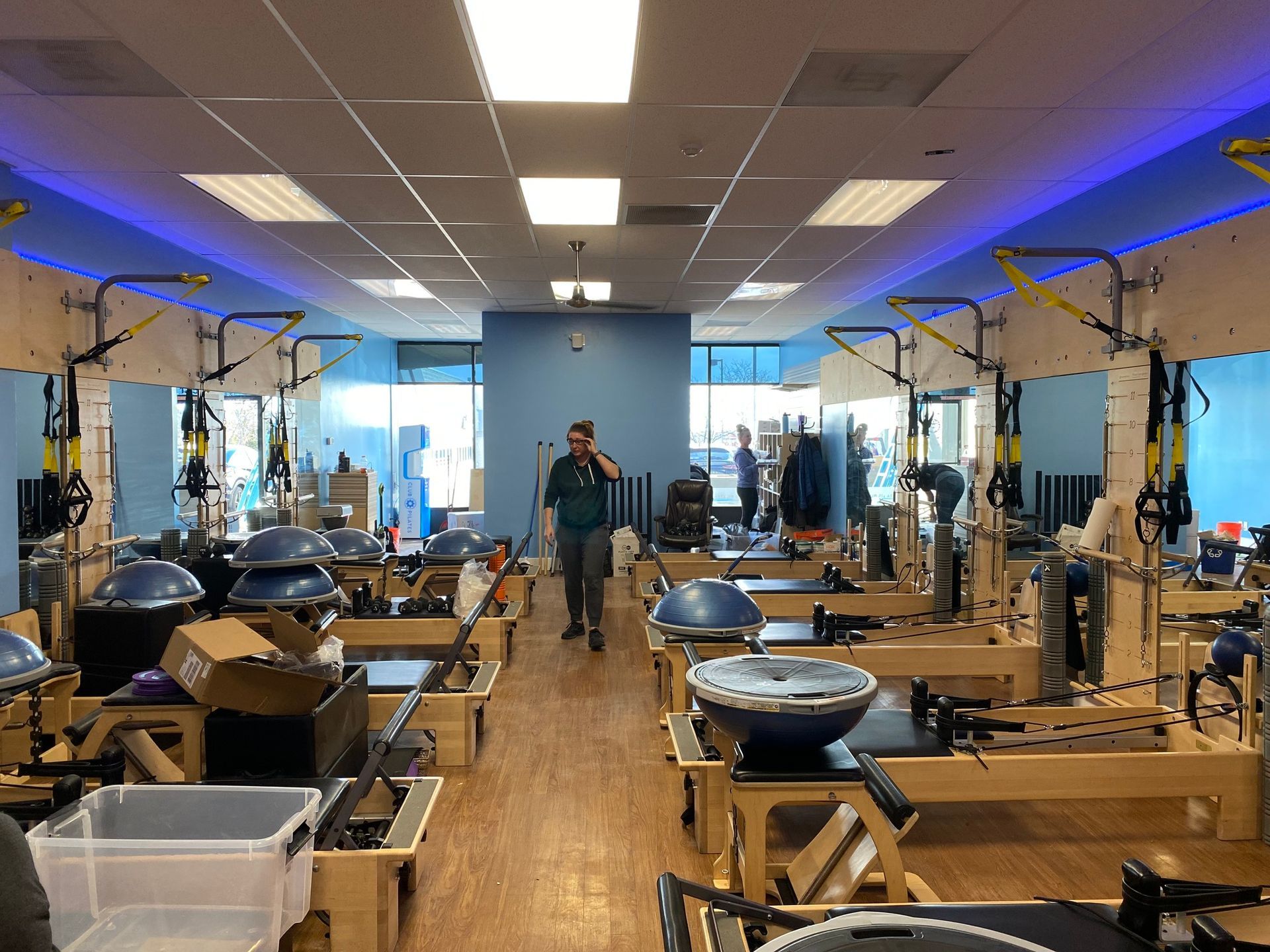 A studio with rows of Pilates reformers and BOSU balls, with a person walking down the center aisle.