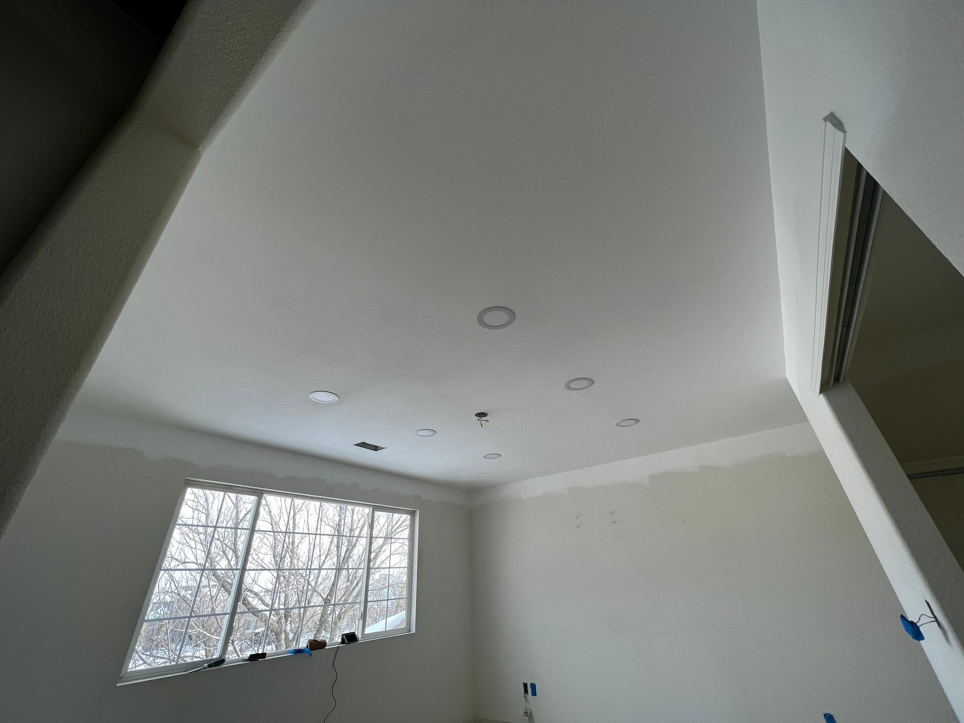 A room under construction with white painted walls and ceiling, recessed lighting holes, and a window looking outdoors.