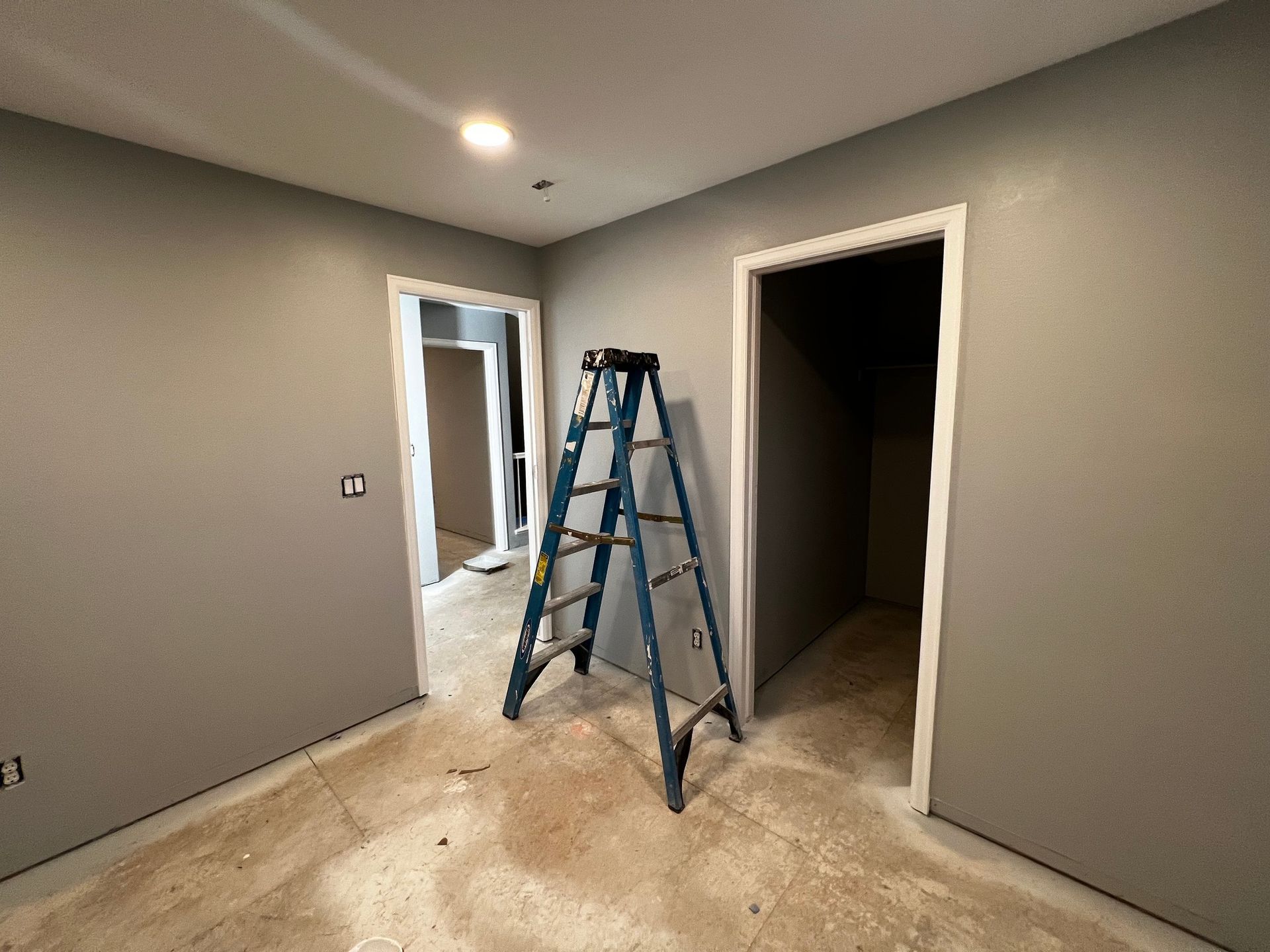 A blue ladder stands in a room with gray walls, two open doorways, and unfinished flooring.