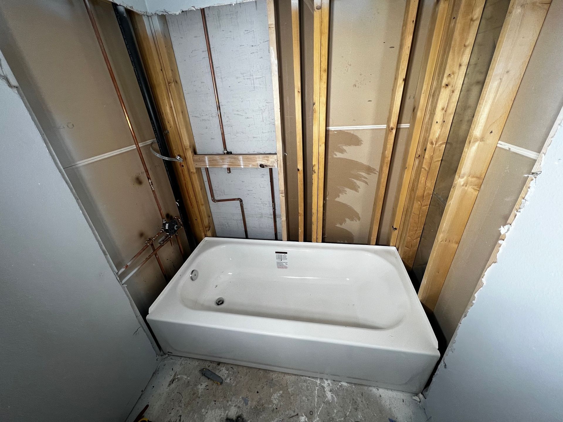 A white bathtub installed in a bathroom alcove with exposed wall studs, wooden framing, and plumbing pipes.