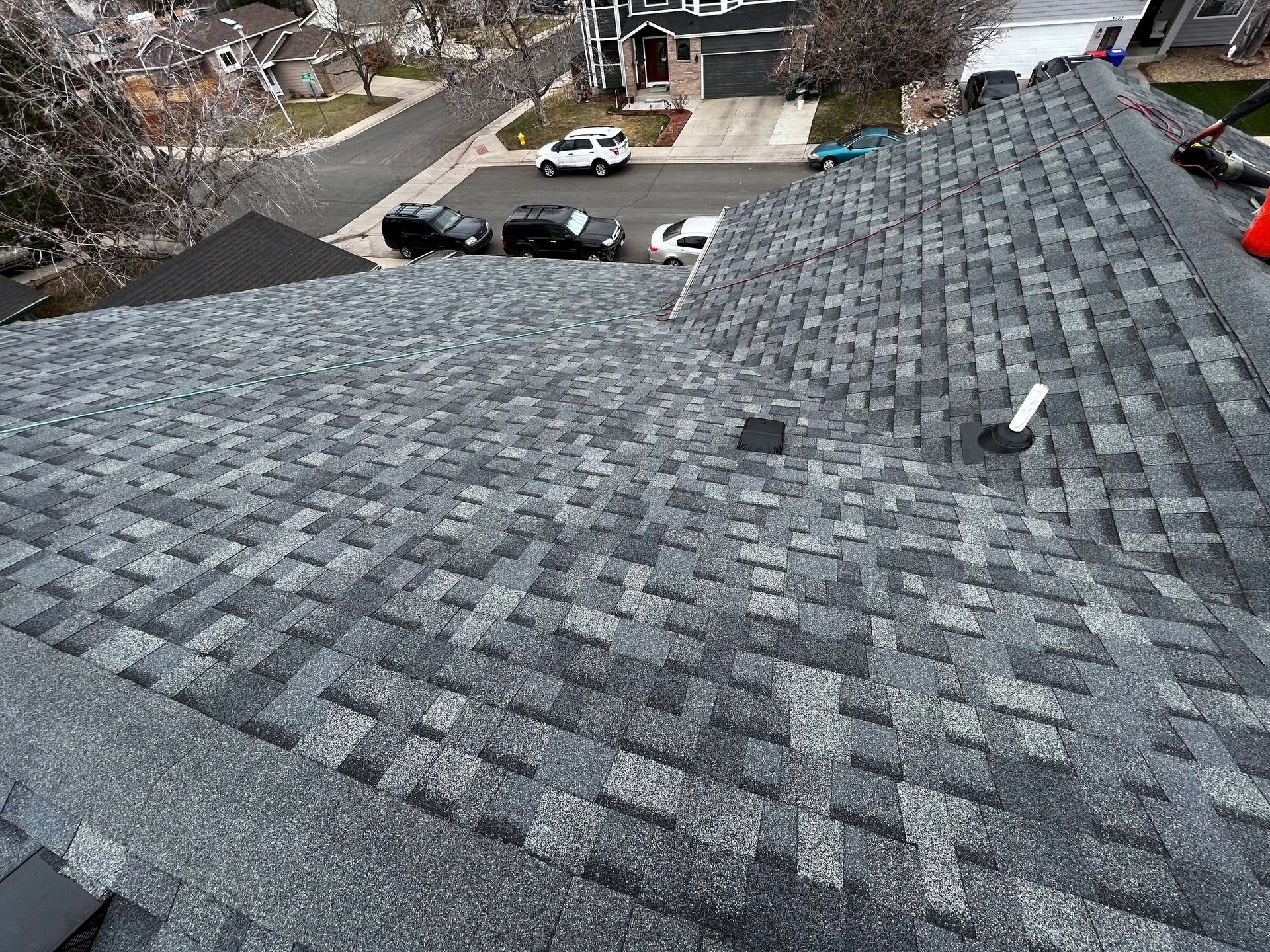 A high-angle view of a grey architectural shingle roof with a vent pipe, overlooking a residential street with cars.