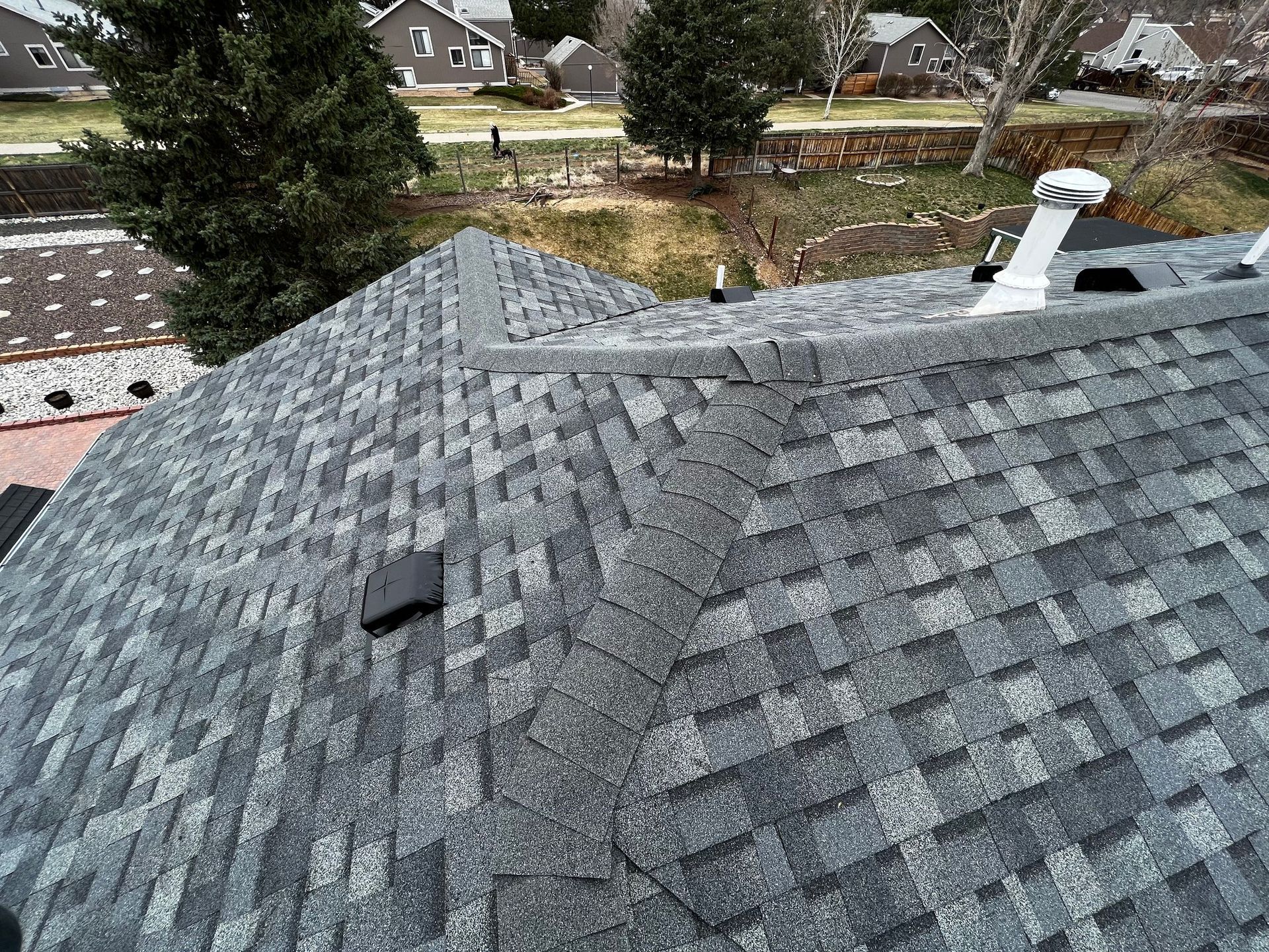 An elevated view of a residential roof featuring gray asphalt shingles, a ridge vent, a plumbing stack, and a roof vent.