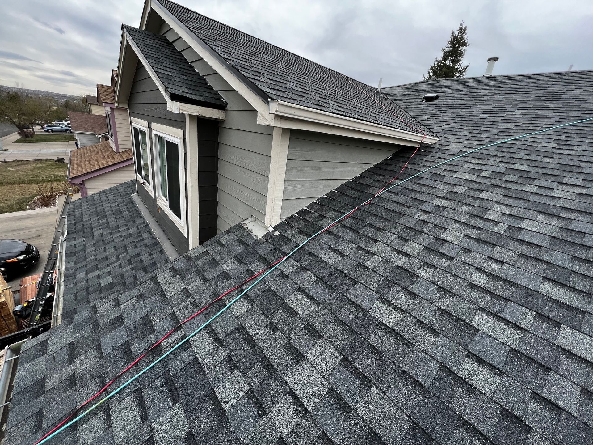High-angle view of a residential shingled roof with a gabled dormer and light-gray siding on an overcast day.
