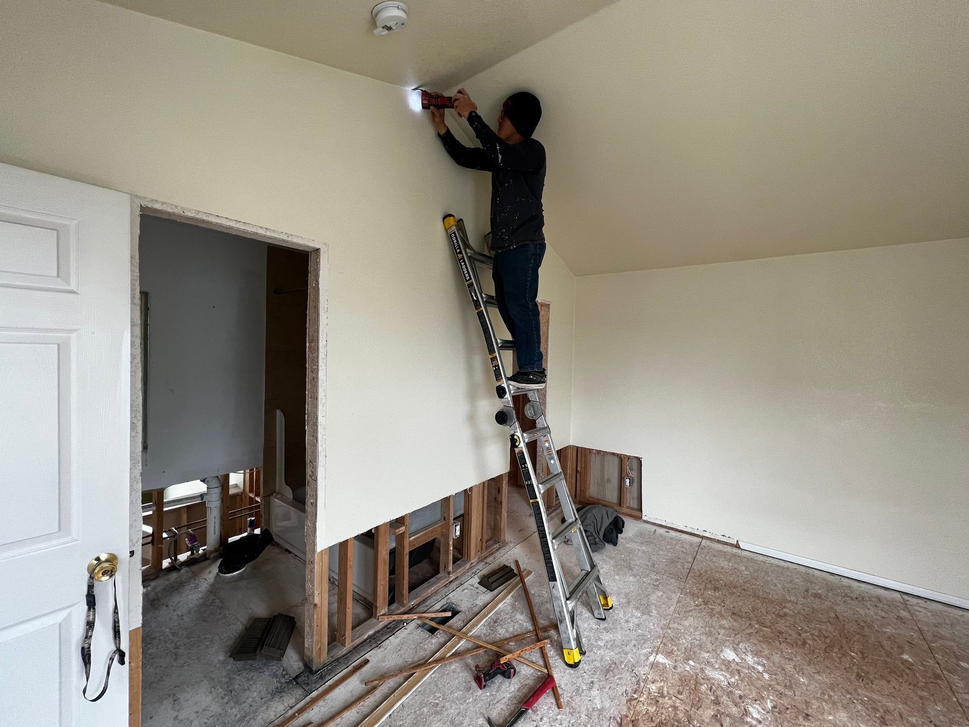 A person stands on a ladder, using a drill on the ceiling of a room under renovation with exposed wall studs.