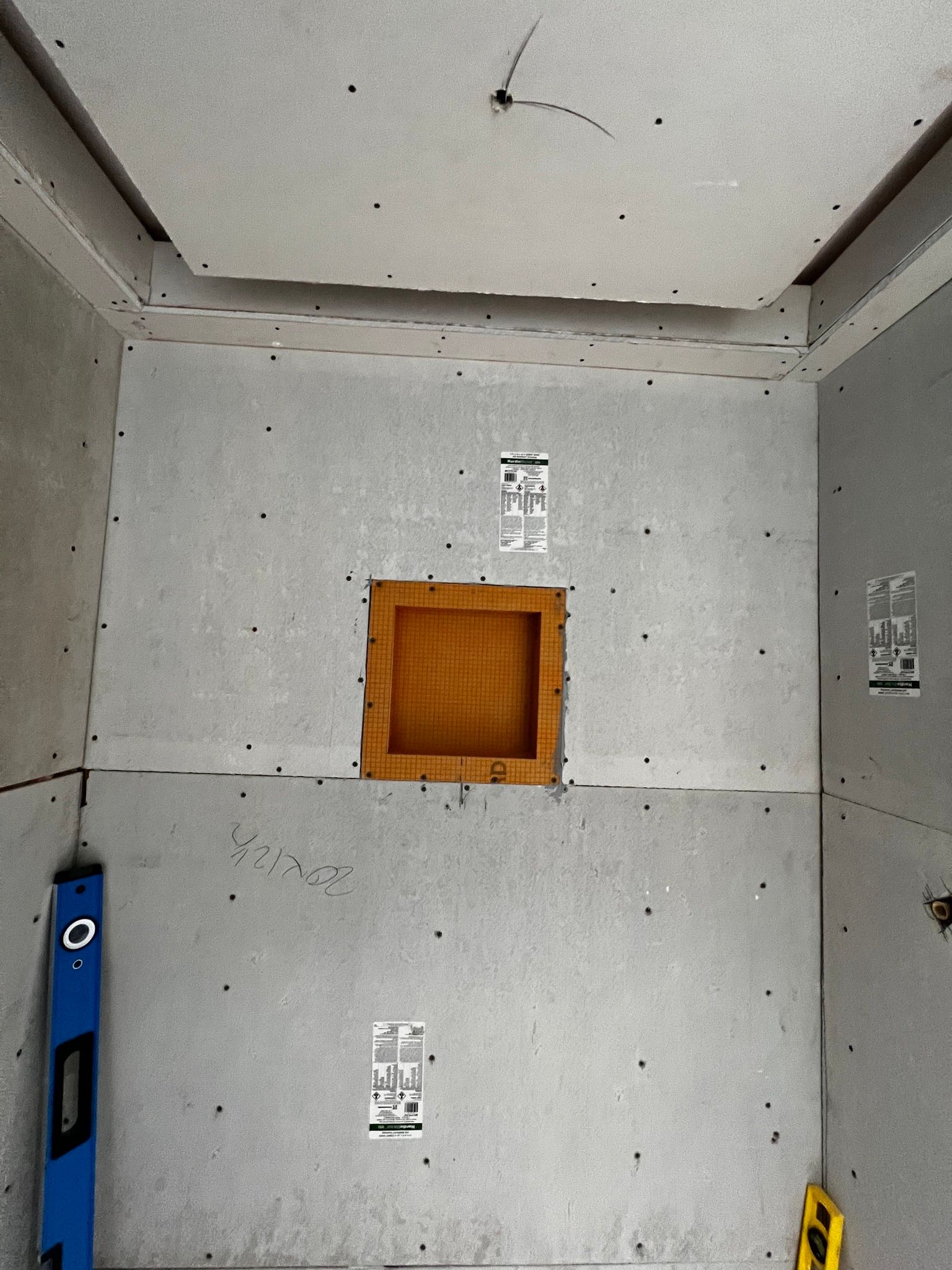 A shower stall under construction with gray cement board walls, a square wooden niche, and a blue level leaning in corner.