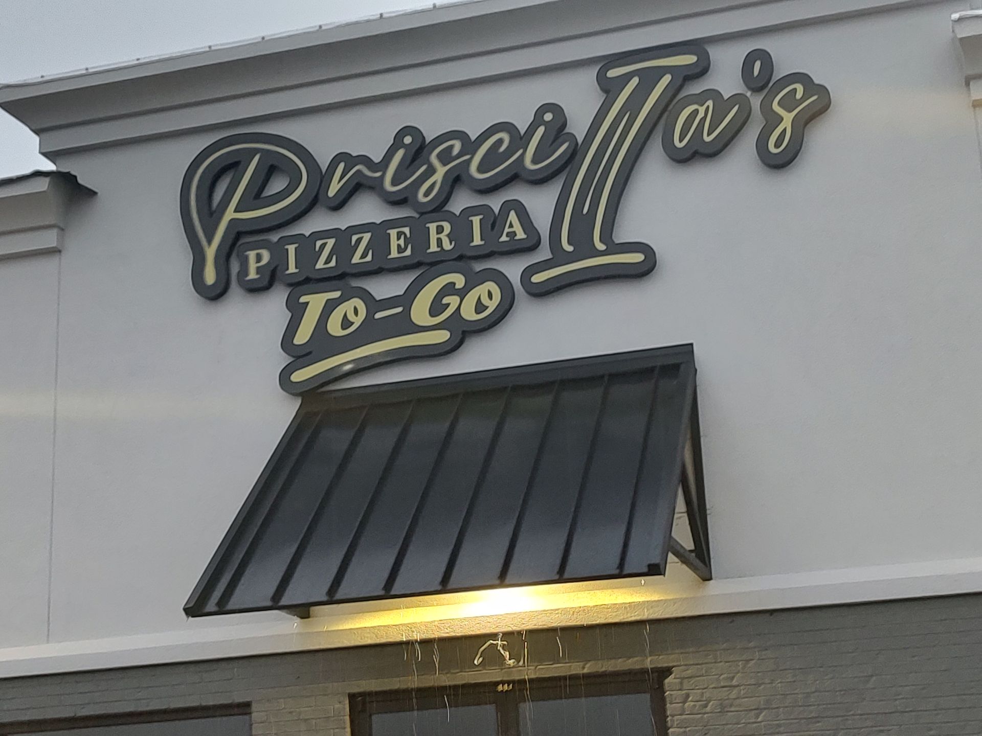 Sign for Priscila's Pizzeria To-Go, dark letters on a light building, awning below.