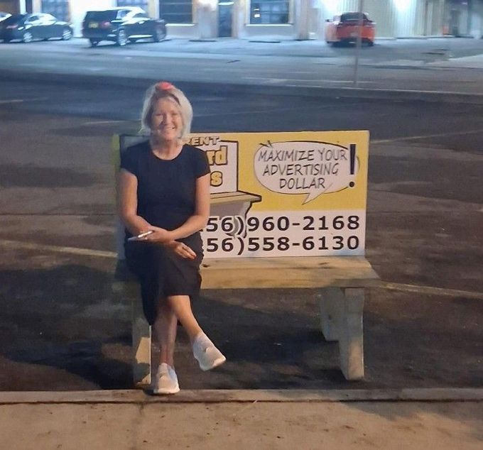 Woman sitting on a bench beside a sign advertising “Maximize your advertising dollars” and phone numbers at night