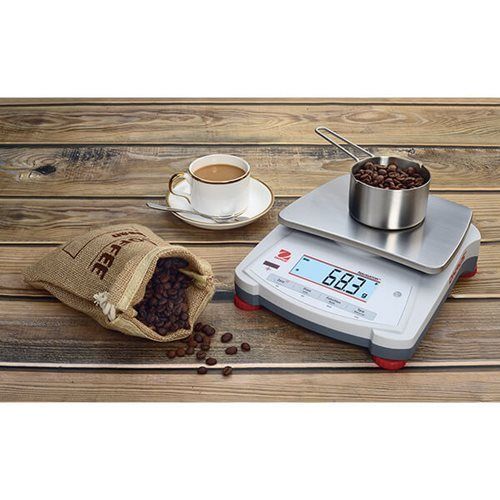 Coffee beans being weighed on a digital scale; cup of coffee, burlap bag, and metal measuring cup on wooden table.
