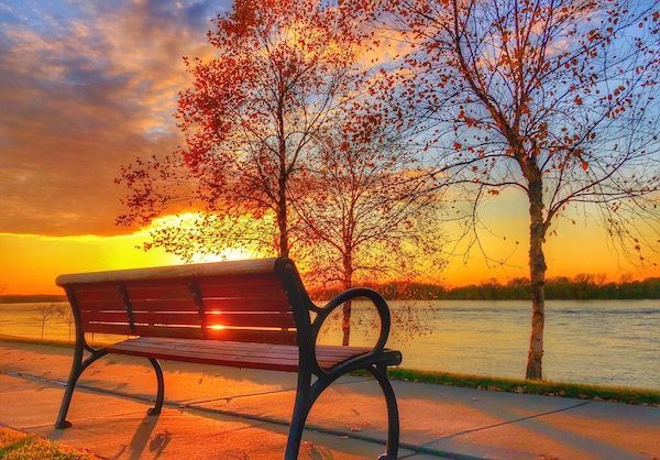 A park bench is sitting on a sidewalk next to a body of water at sunset.