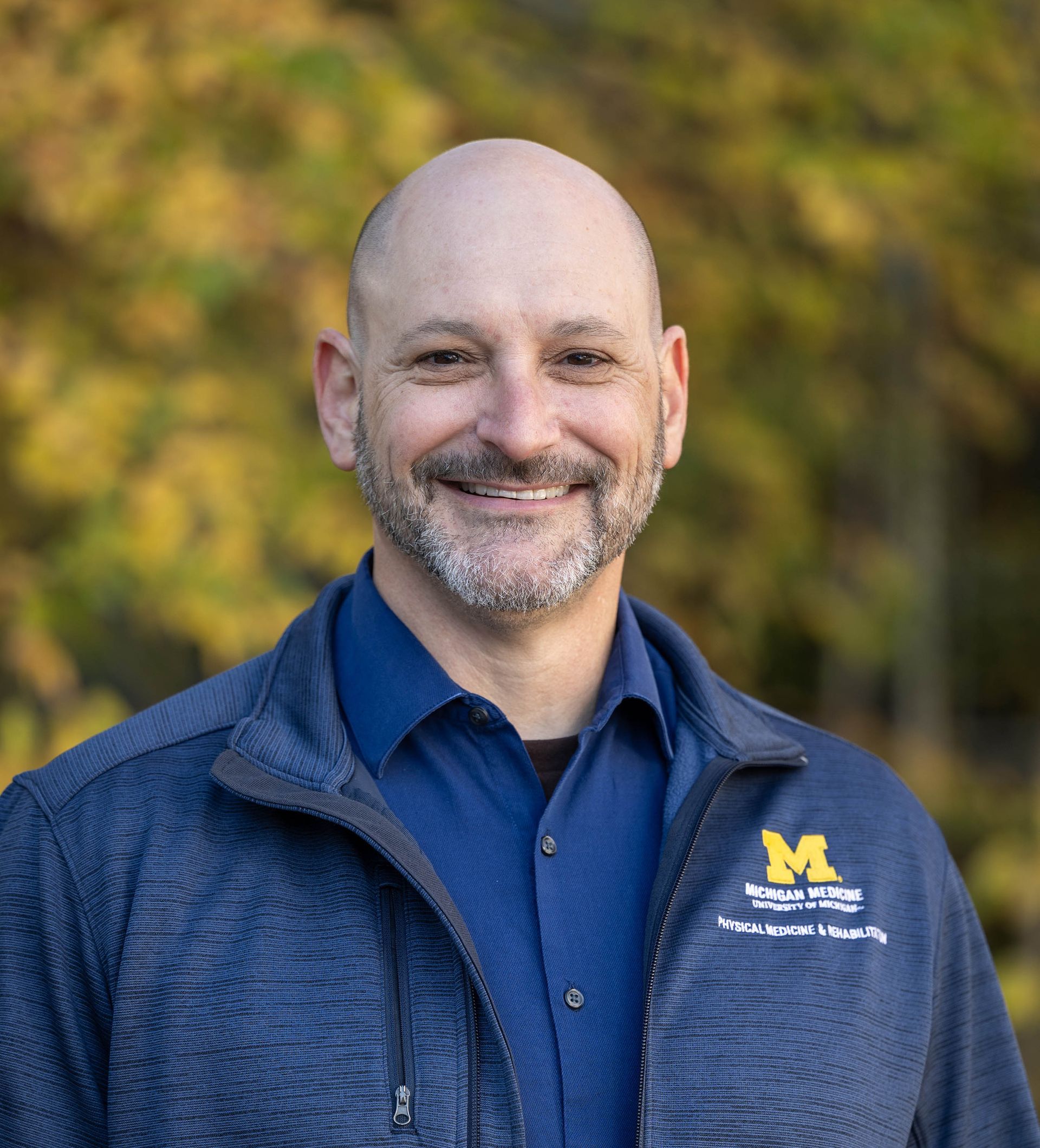 A smiling person wears a blue collared shirt and a navy zip-up jacket with a University of Michigan logo outdoors.