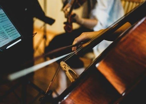 A cellist with bow and cello, playing in an orchestra. Other musicians visible in the background.