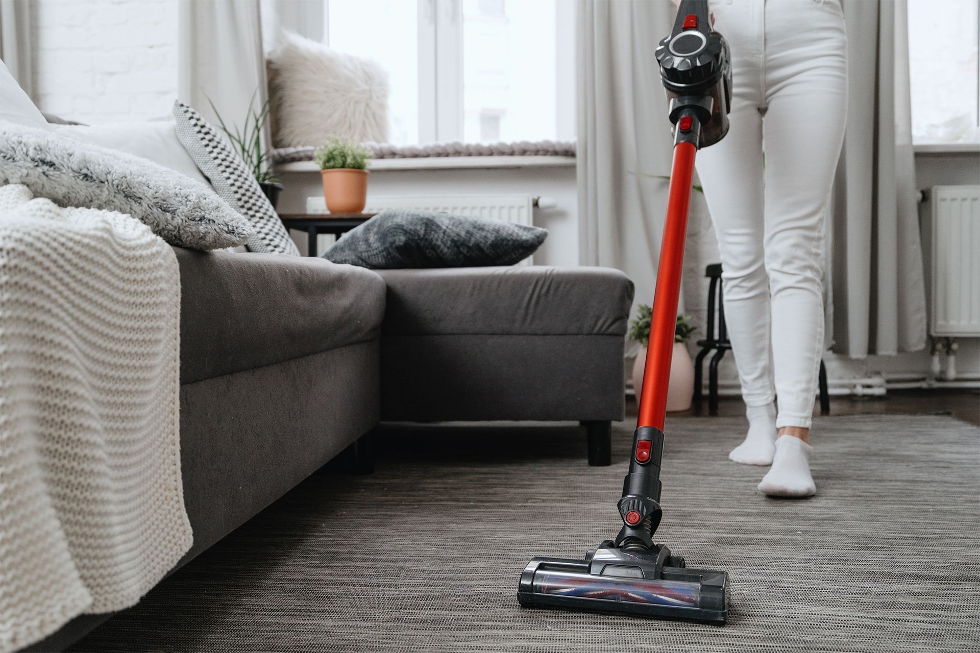 Woman in white jeans vacuums a gray rug with a red stick vacuum in a living room.