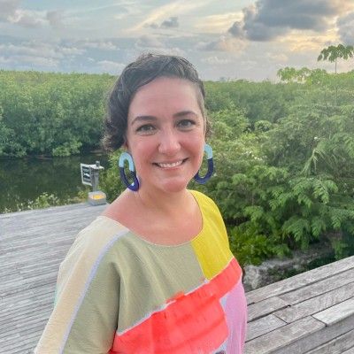 Smiling woman in a colorful top standing on a wooden deck by lush greenery at sunset