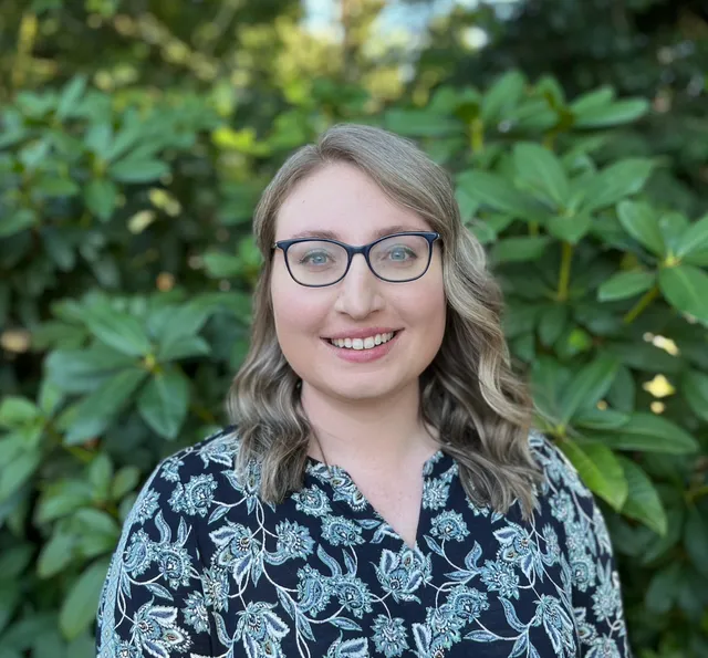 Smiling person with glasses in a patterned blouse, standing before leafy green shrubs.